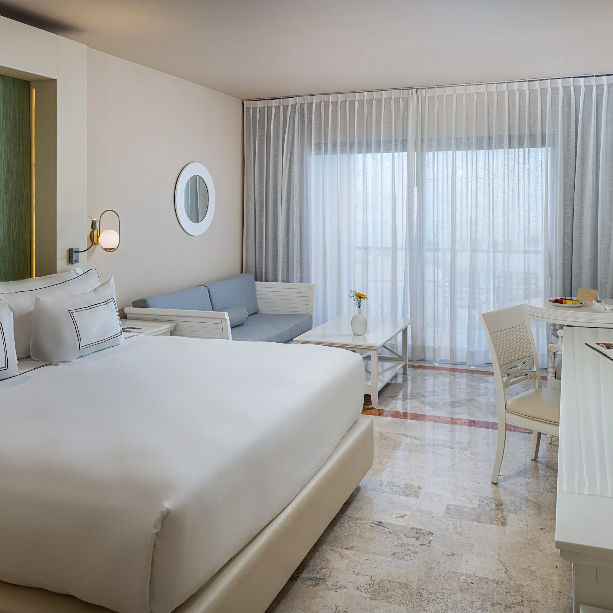 Spacious modern hotel room with king bed, sofa, and dining area by bright window.