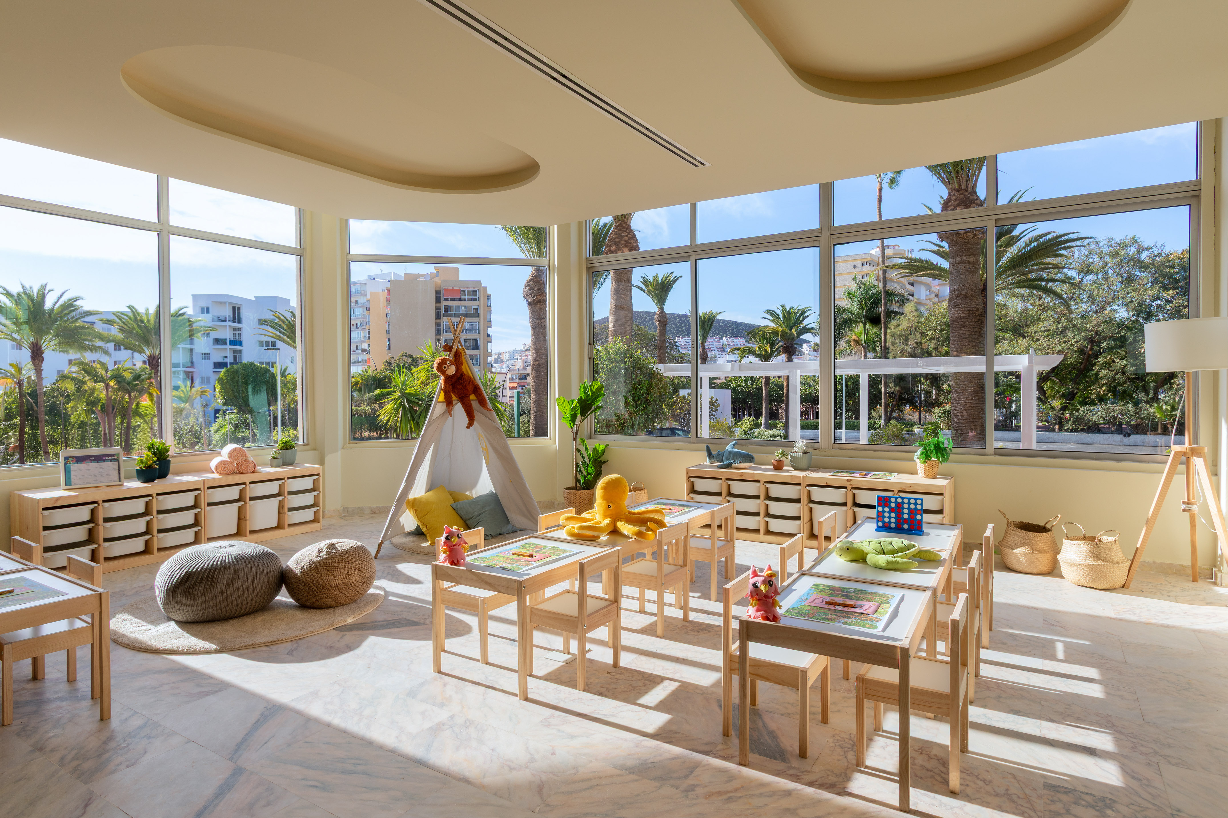 Bright children's playroom with tables, teepee, storage, and tropical window views.