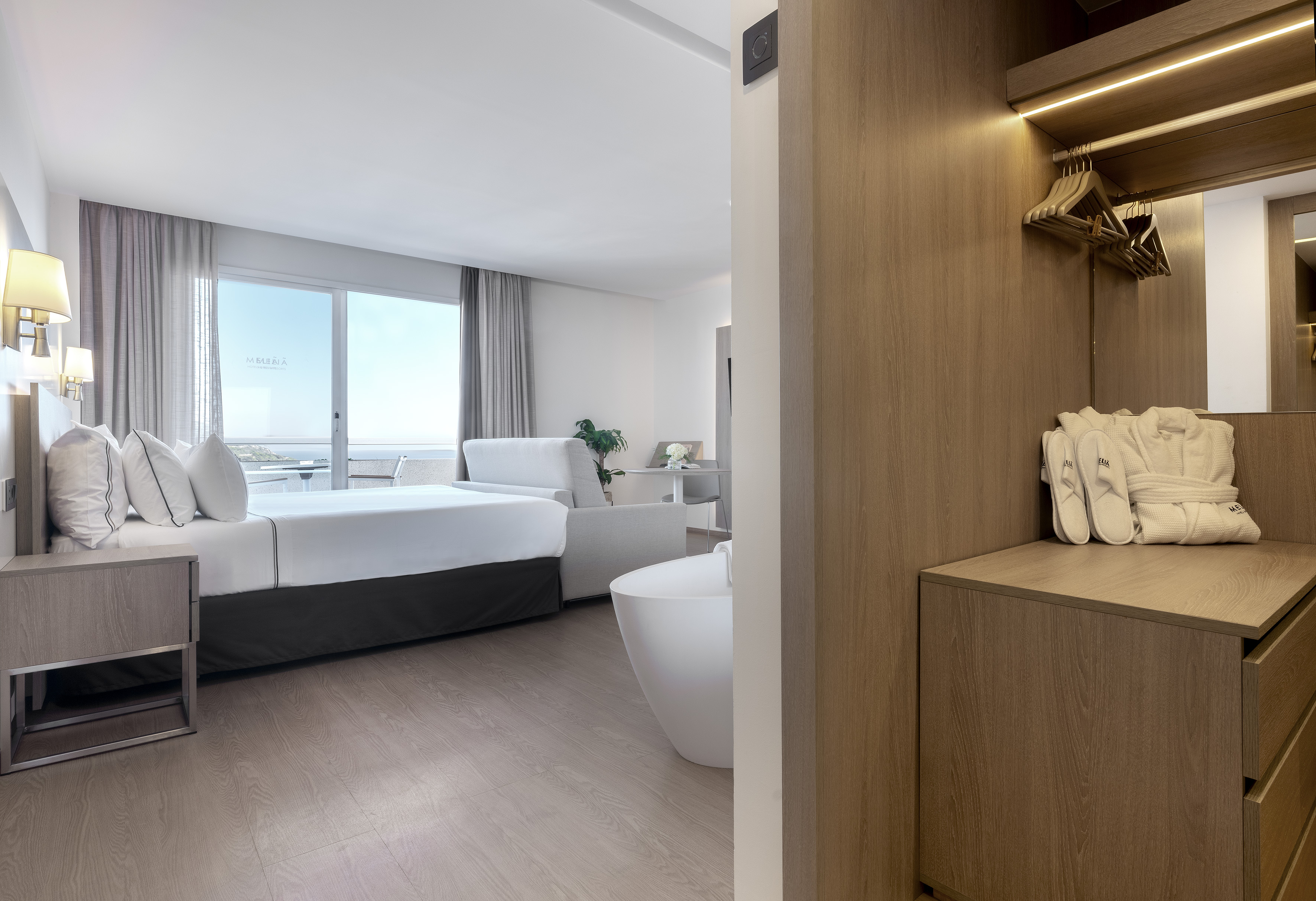 Bright hotel suite: bed, sea view balcony, in-room tub, open wardrobe.