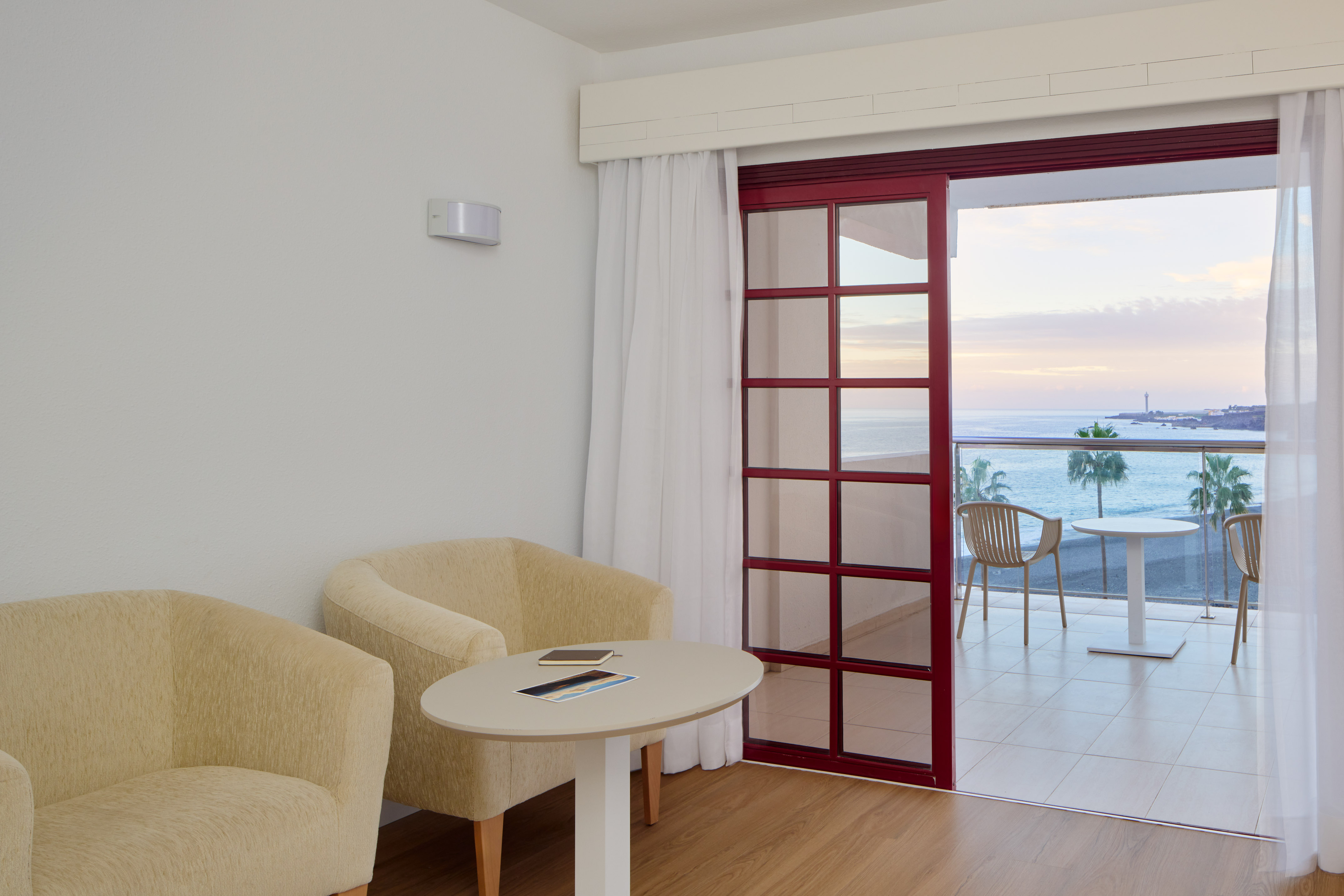 Coastal hotel room featuring balcony with sea view, palm trees, and lighthouse.