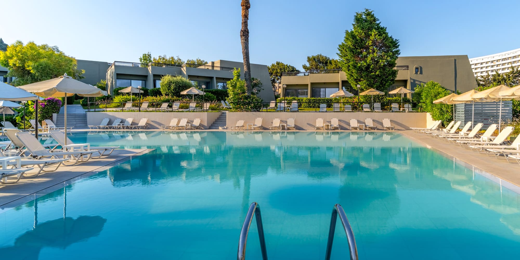 Sunny hotel resort swimming pool with loungers, umbrellas, and palm trees.