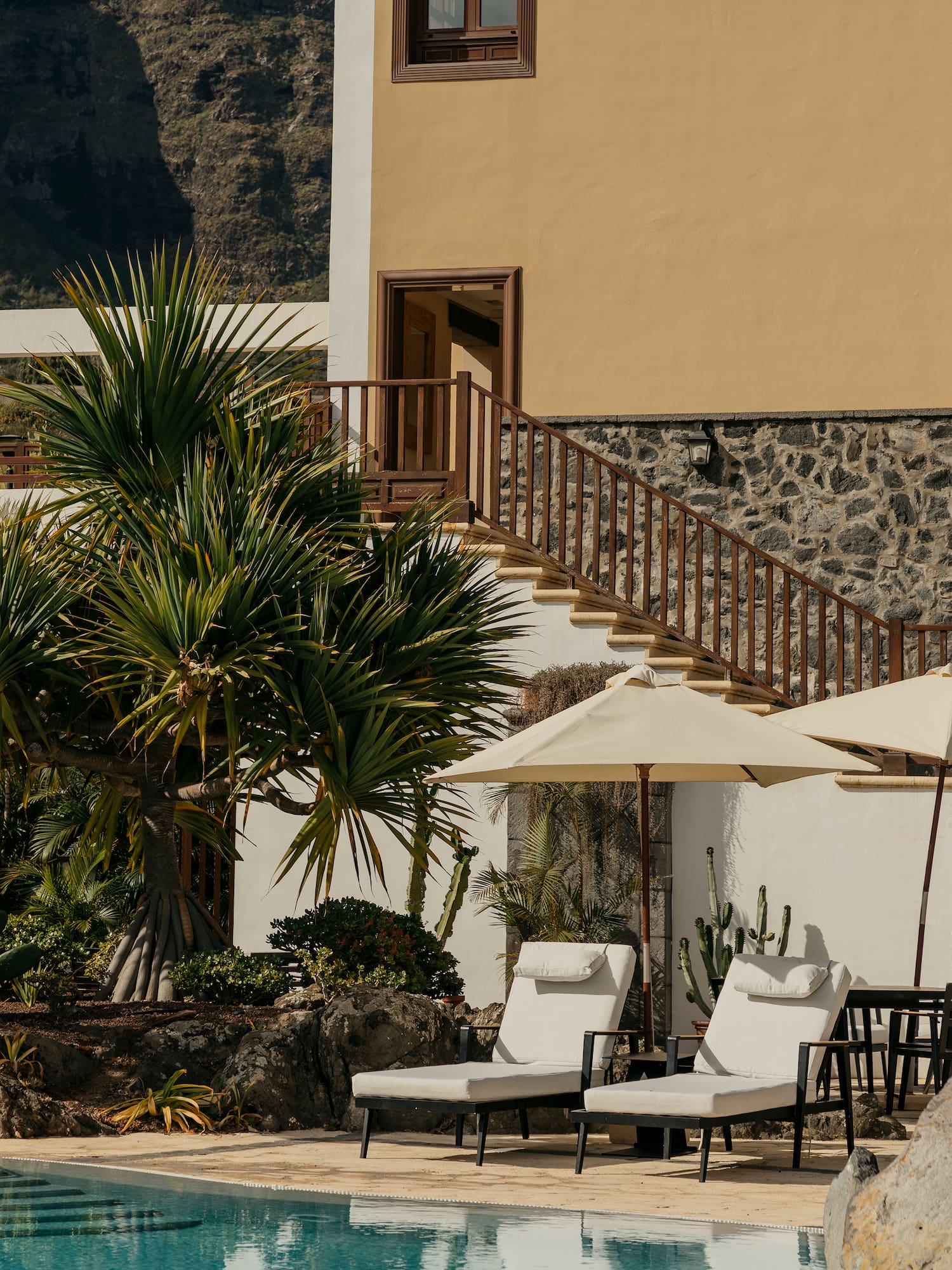 Sunny resort pool with lounge chairs, palm trees, yellow building, mountain view.