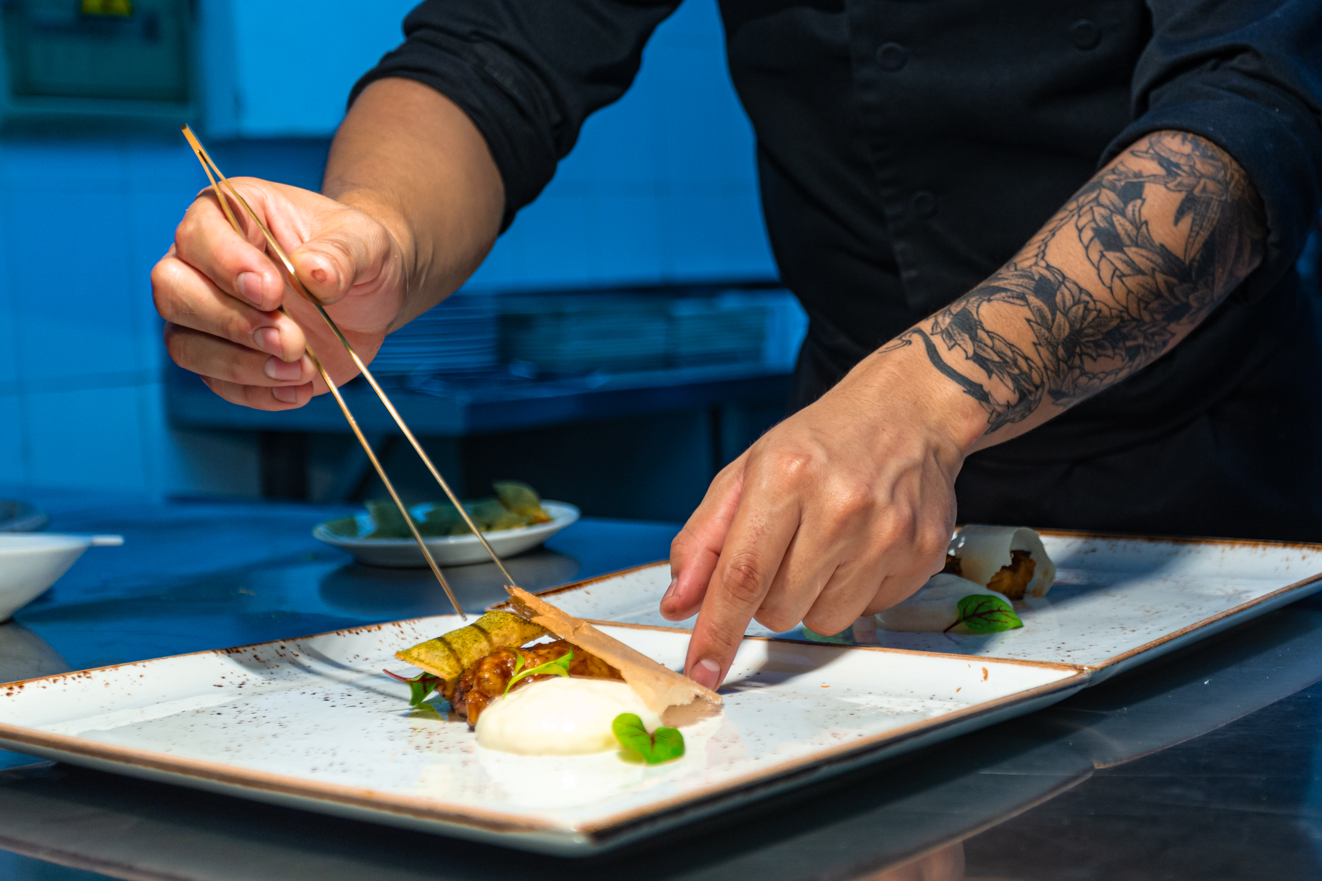 Chef with tattooed arm plating gourmet food with precision tweezers.