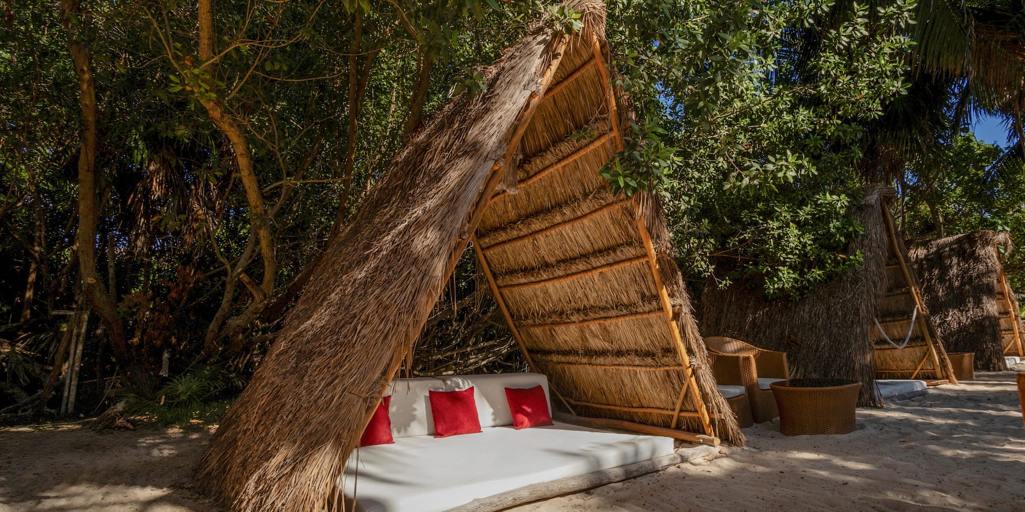 Tropical thatched cabana daybed with red pillows on sandy beach.