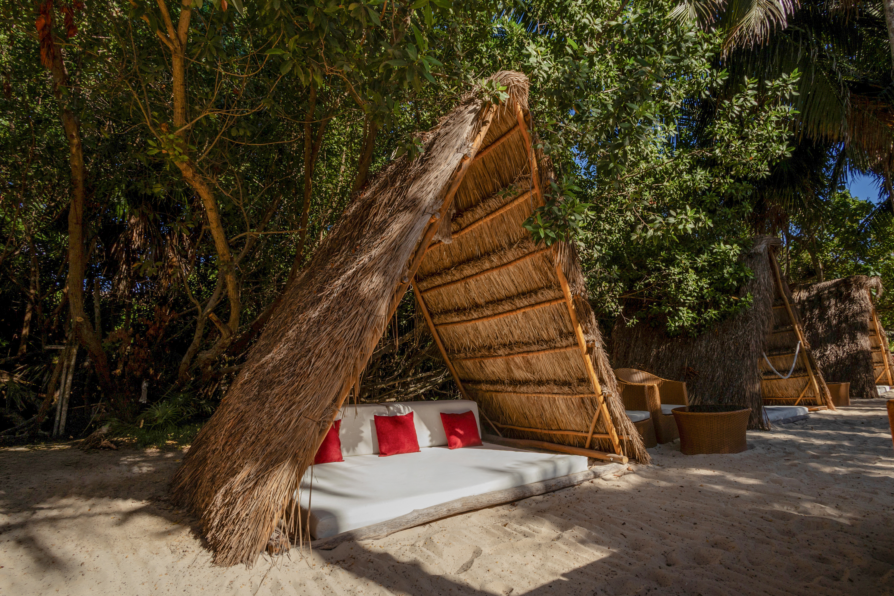 Tropical thatched cabana daybed with red pillows on sandy beach.