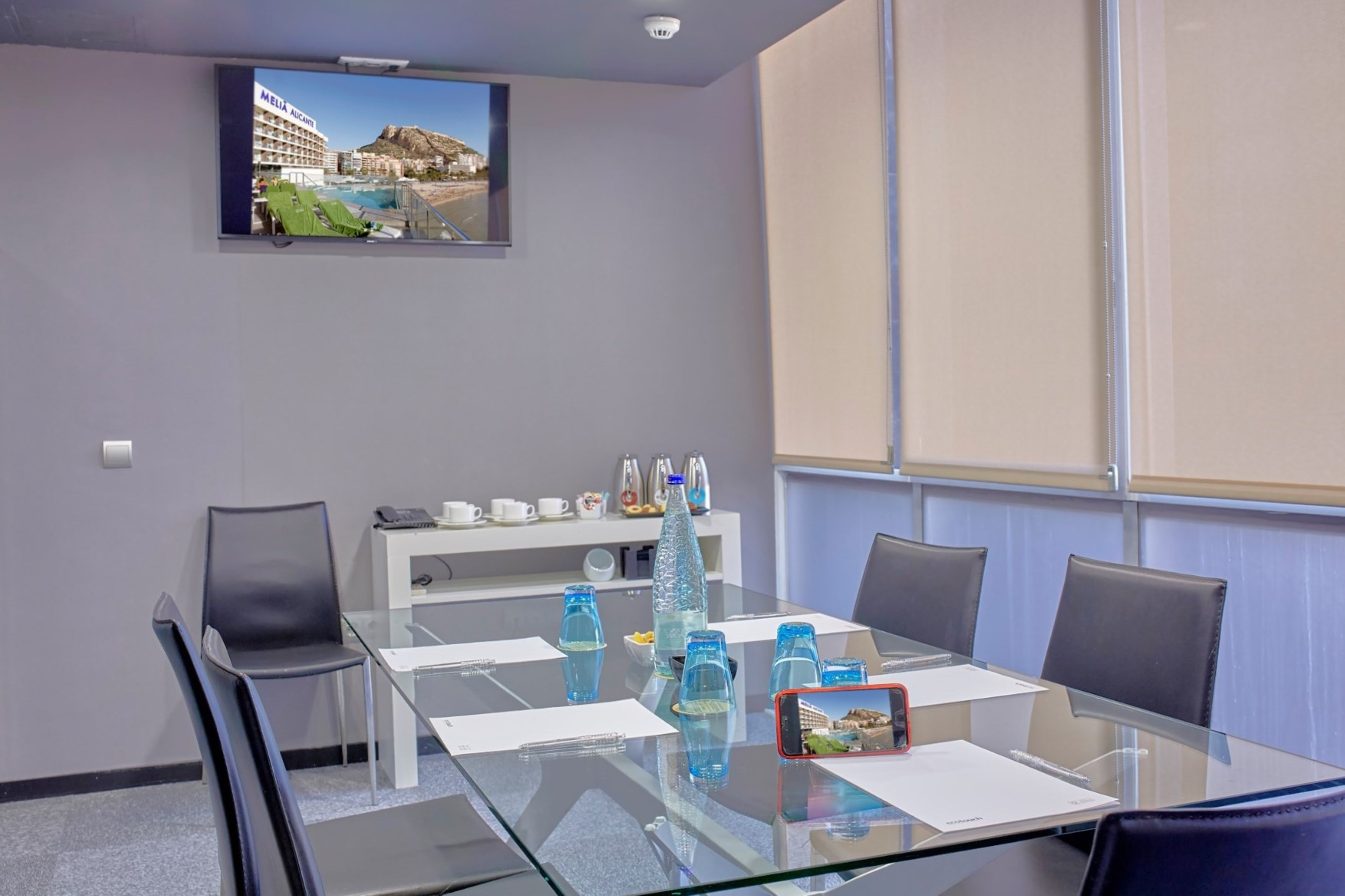 Modern hotel meeting room with glass table, black chairs, and large TV screen.