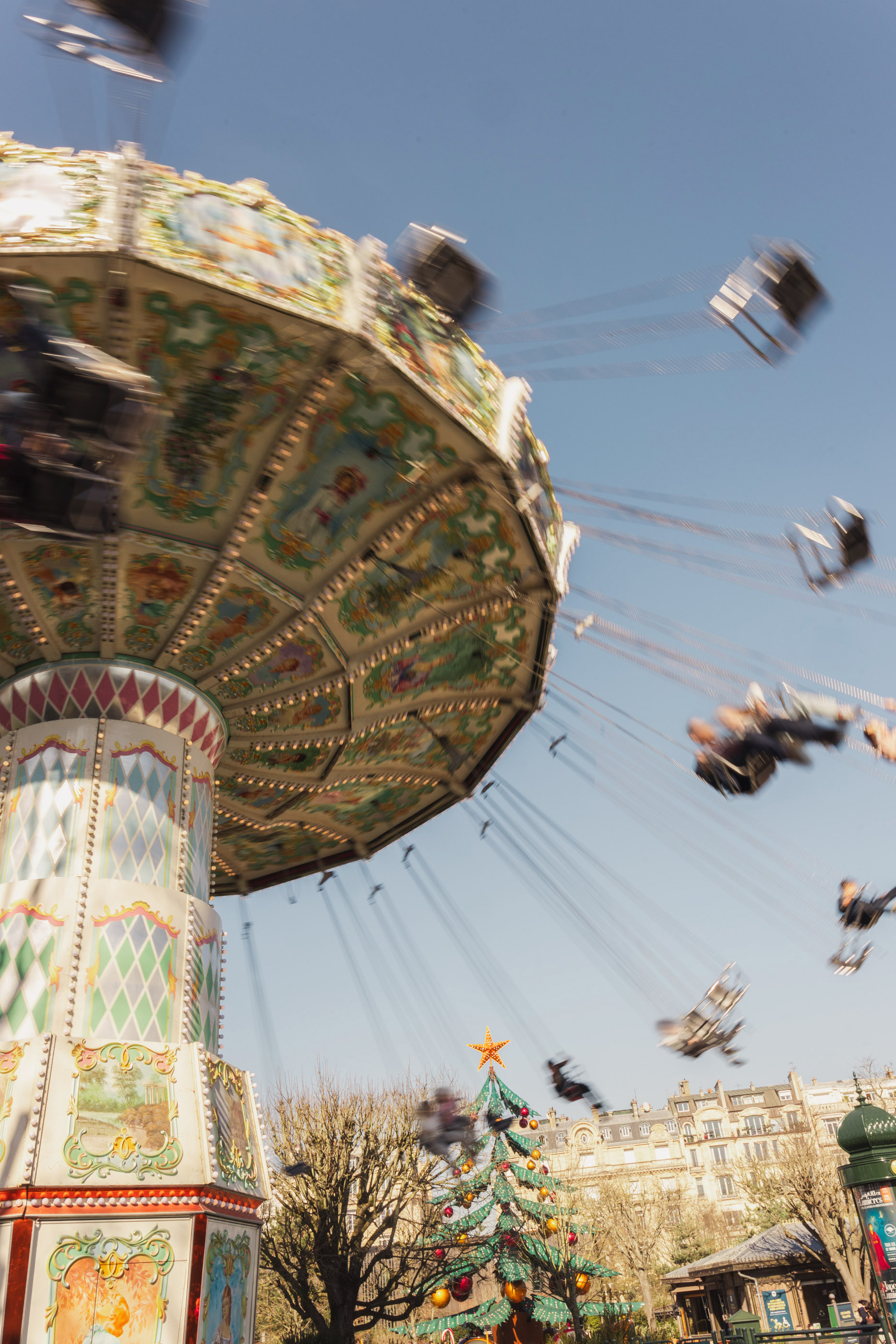 Motion-blurred swing ride with riders against blue sky and Christmas tree.