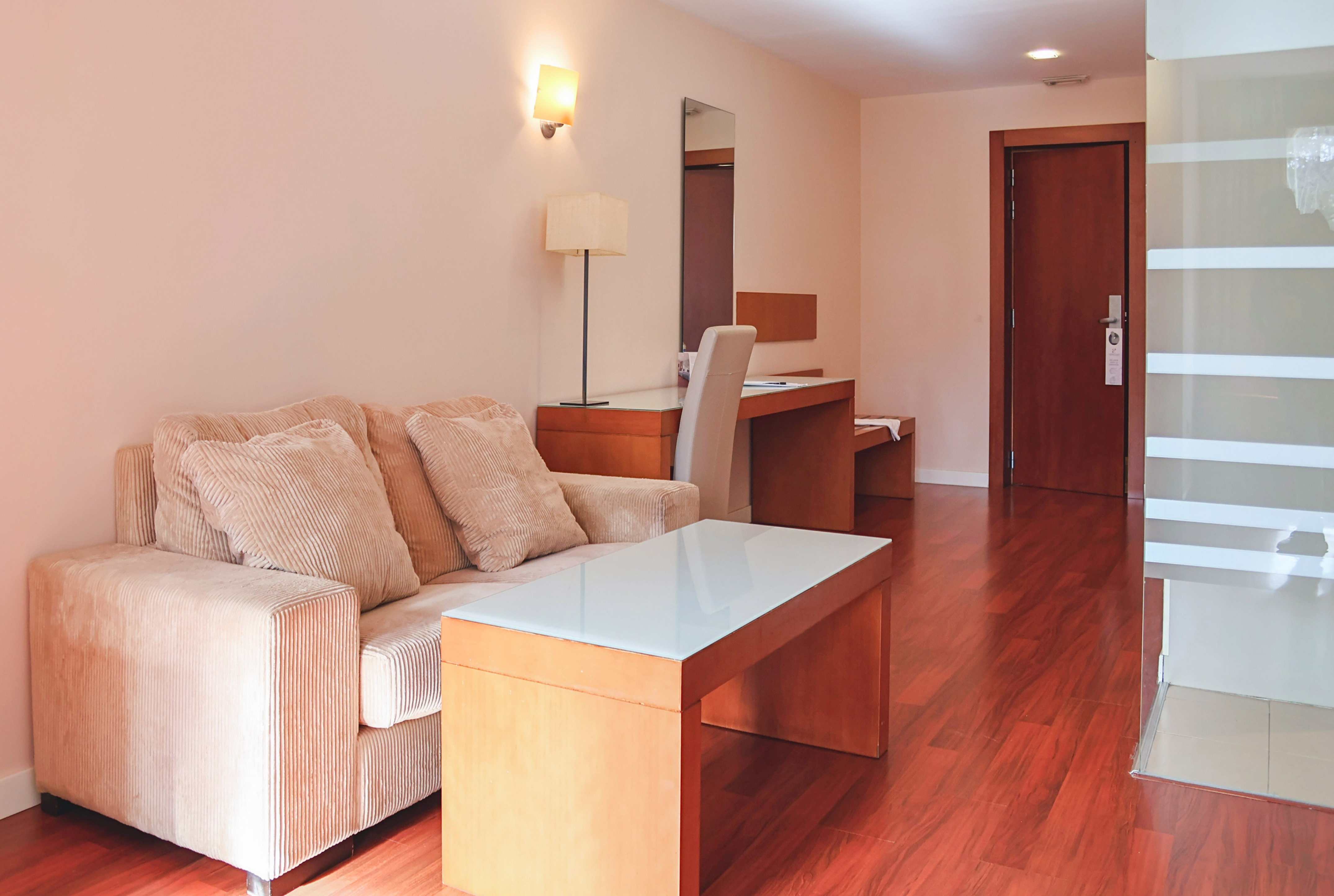 Modern hotel room with beige sofa, wooden desk, and hardwood floor.