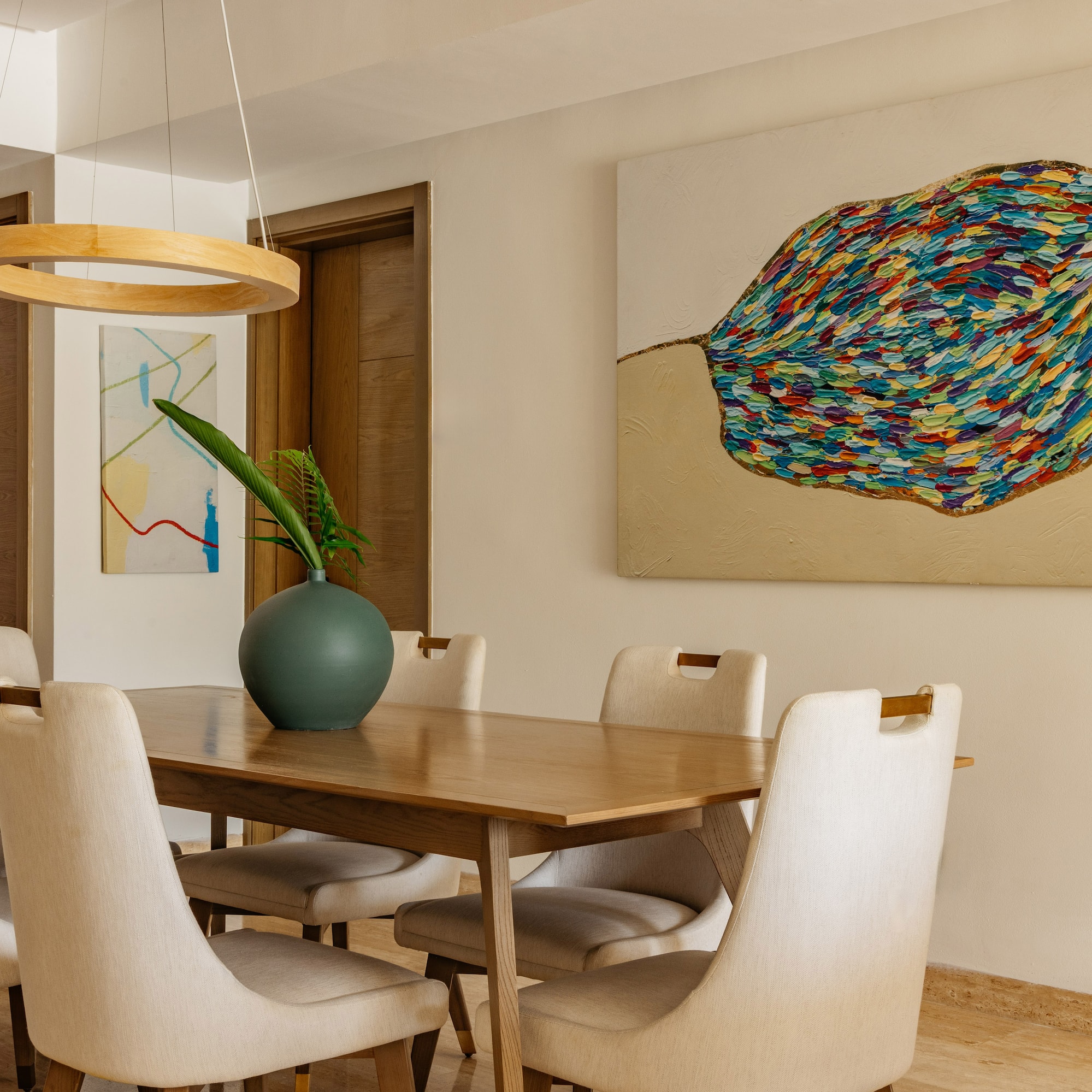 Modern dining room: wooden table, cream chairs, vibrant abstract leaf painting.