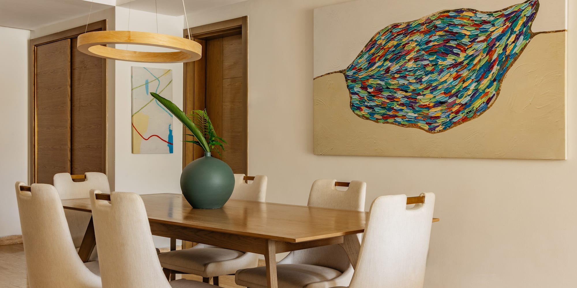 Modern dining room: wooden table, cream chairs, vibrant abstract leaf painting.