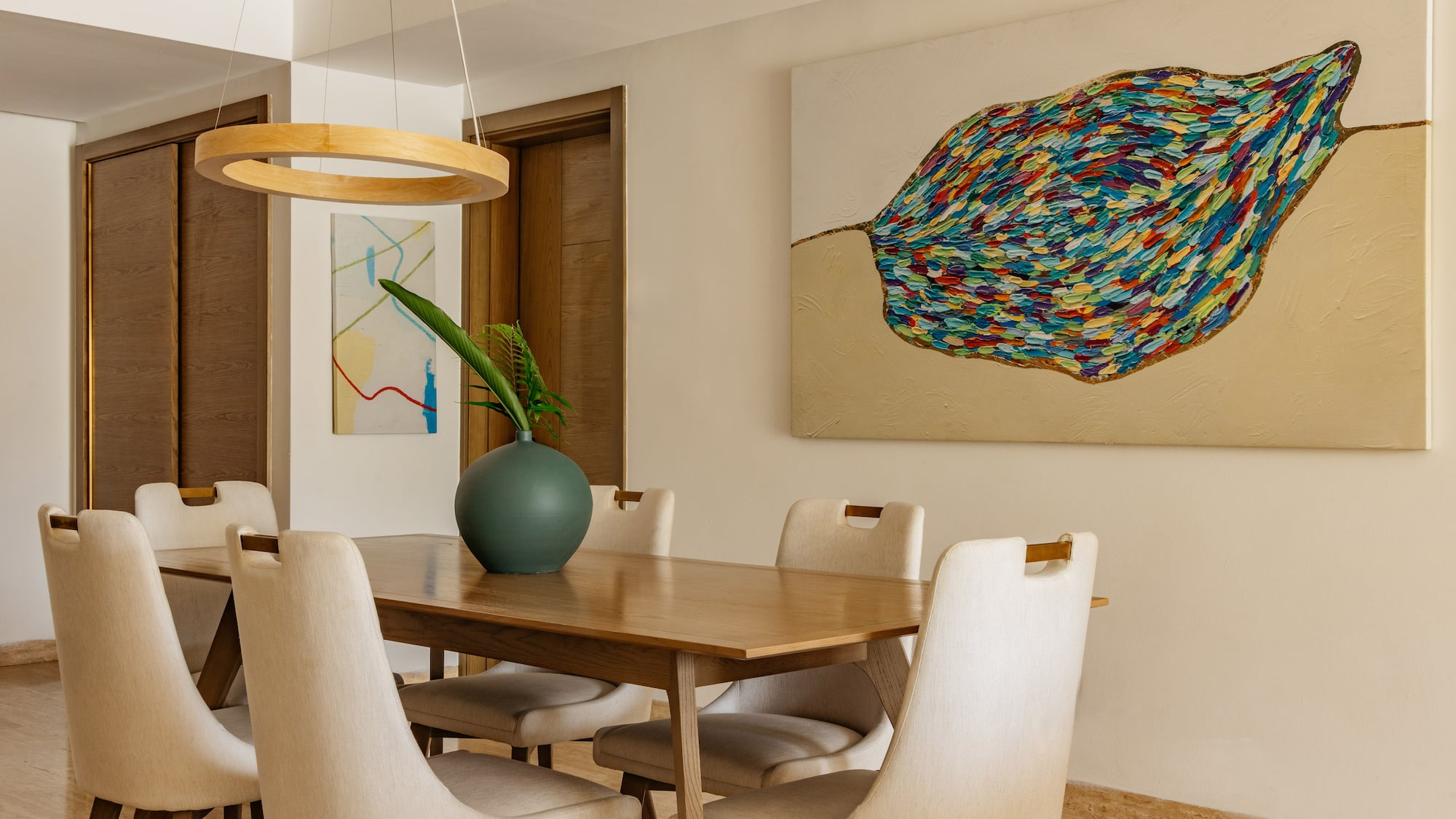 Modern dining room: wooden table, cream chairs, vibrant abstract leaf painting.