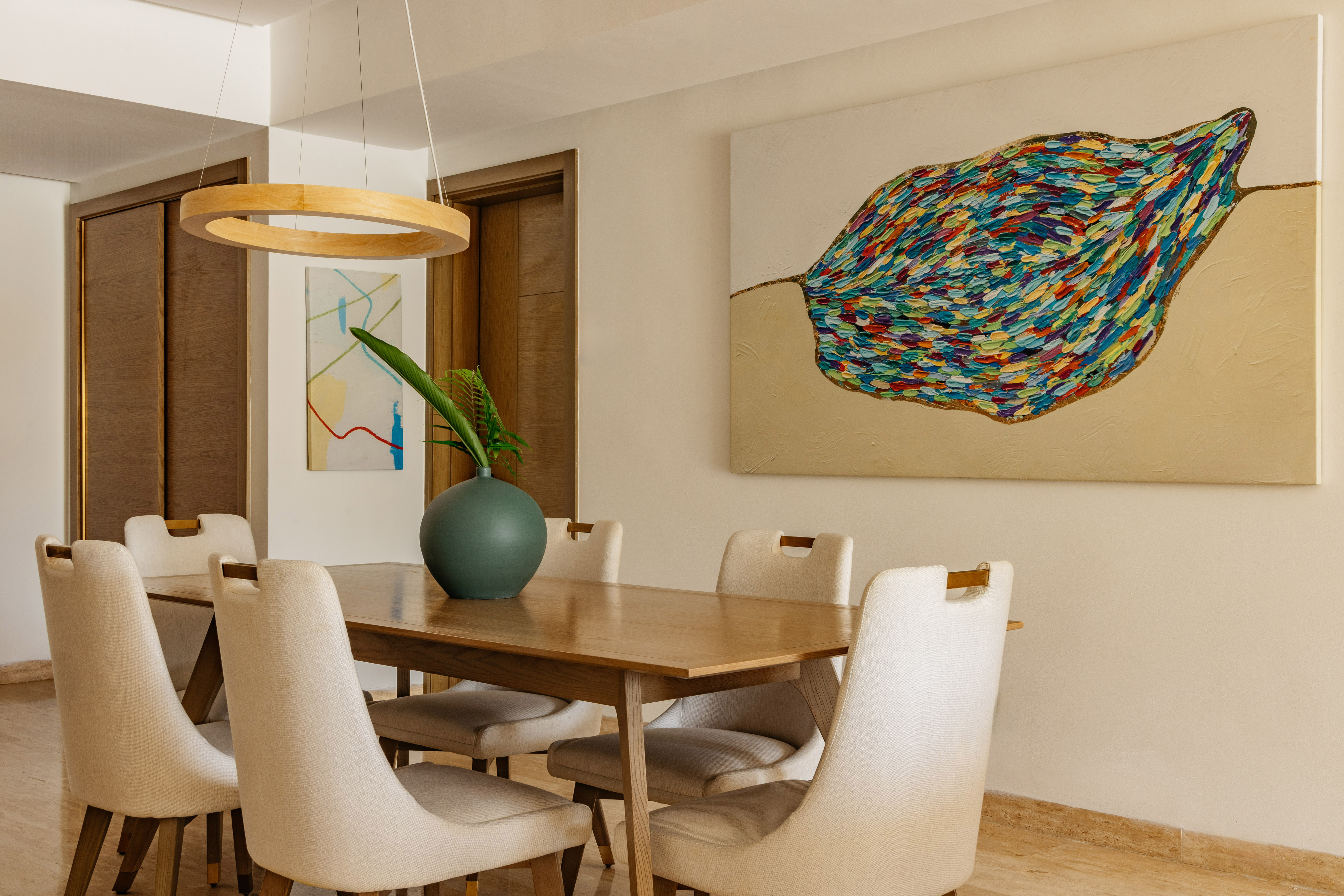 Modern dining room: wooden table, cream chairs, vibrant abstract leaf painting.