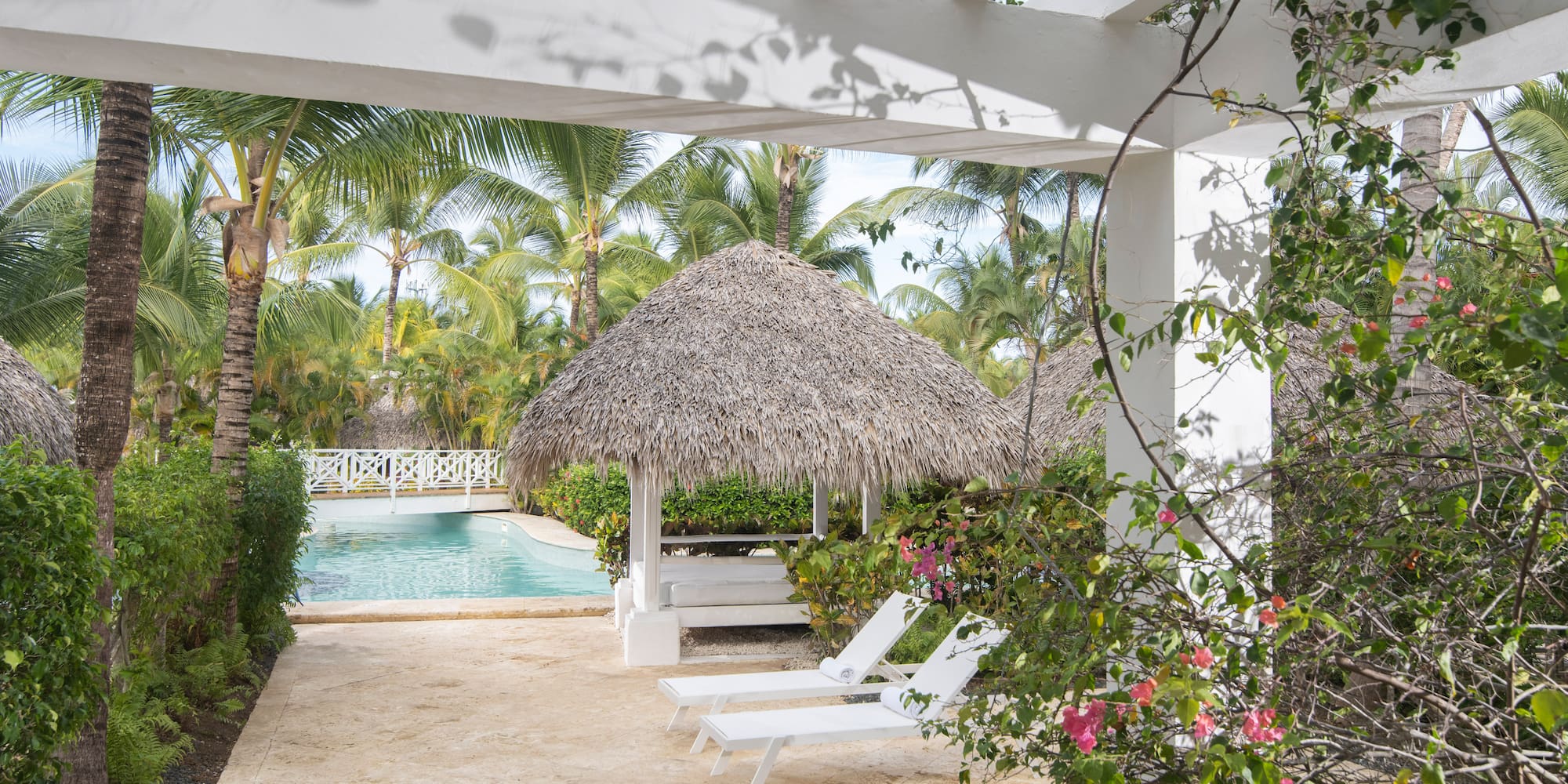 Resort pool area with pergola, thatched gazebo, lounge chairs, and palm trees.