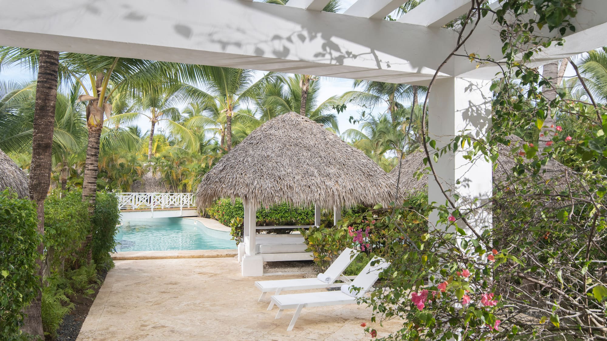 Resort pool area with pergola, thatched gazebo, lounge chairs, and palm trees.