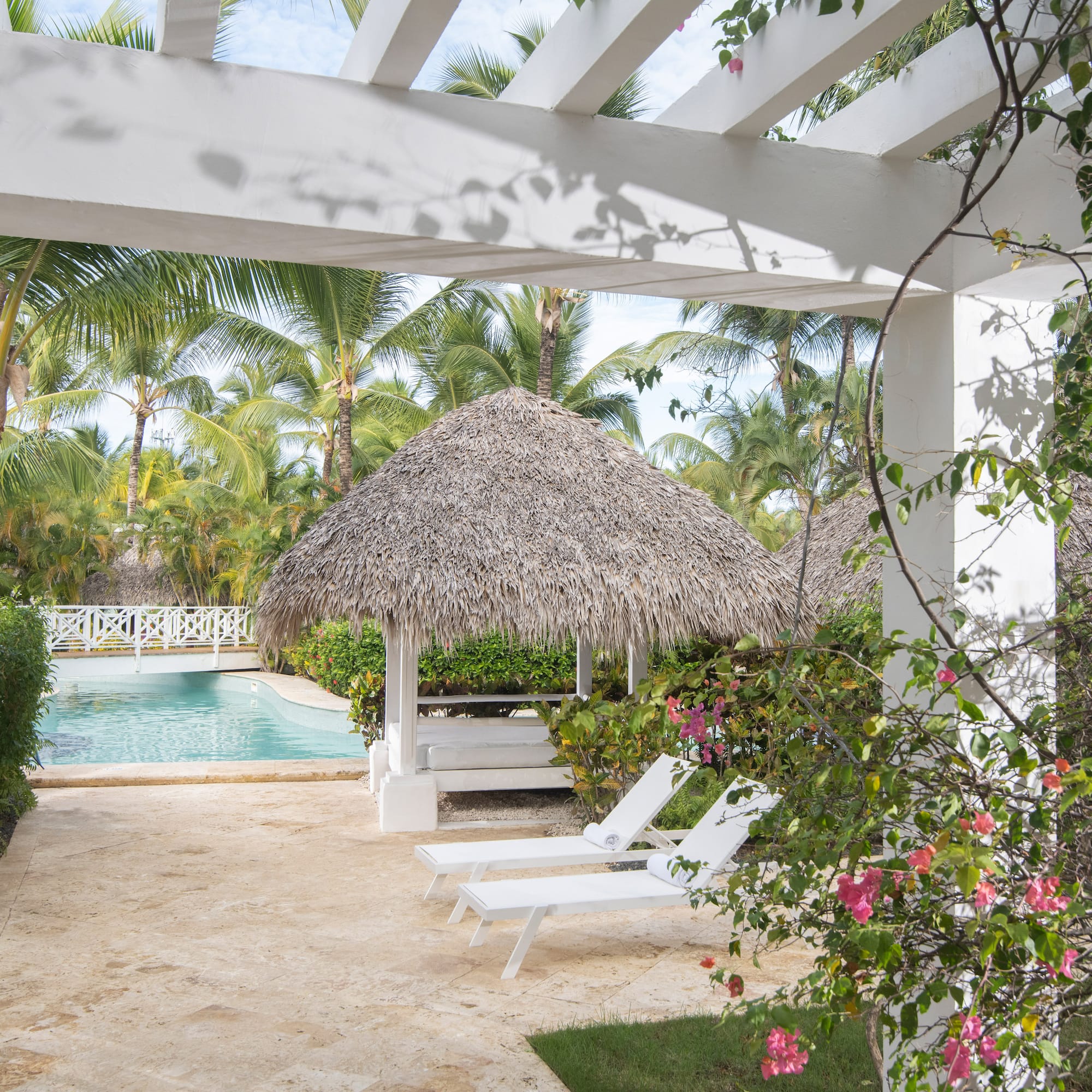 Resort pool area with pergola, thatched gazebo, lounge chairs, and palm trees.