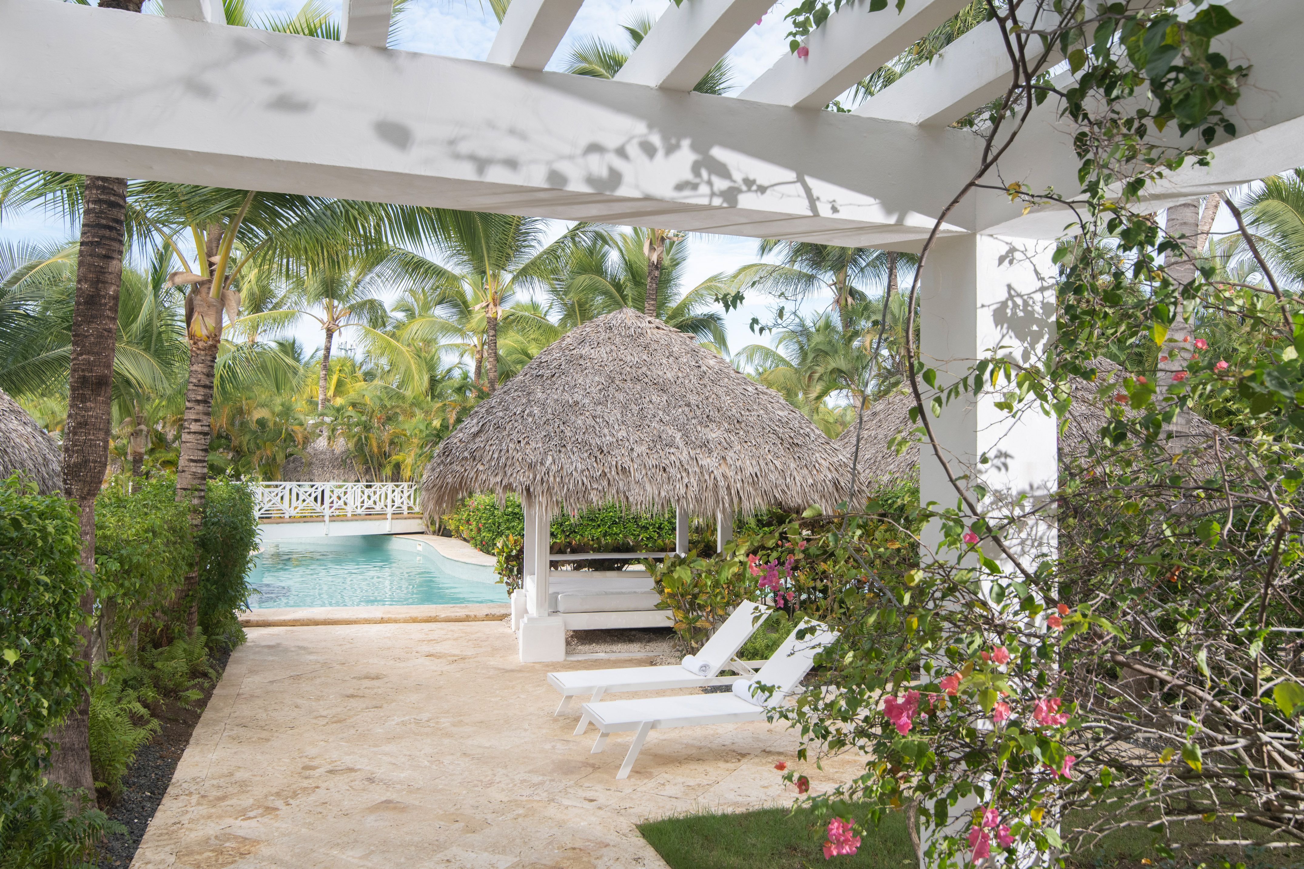 Resort pool area with pergola, thatched gazebo, lounge chairs, and palm trees.
