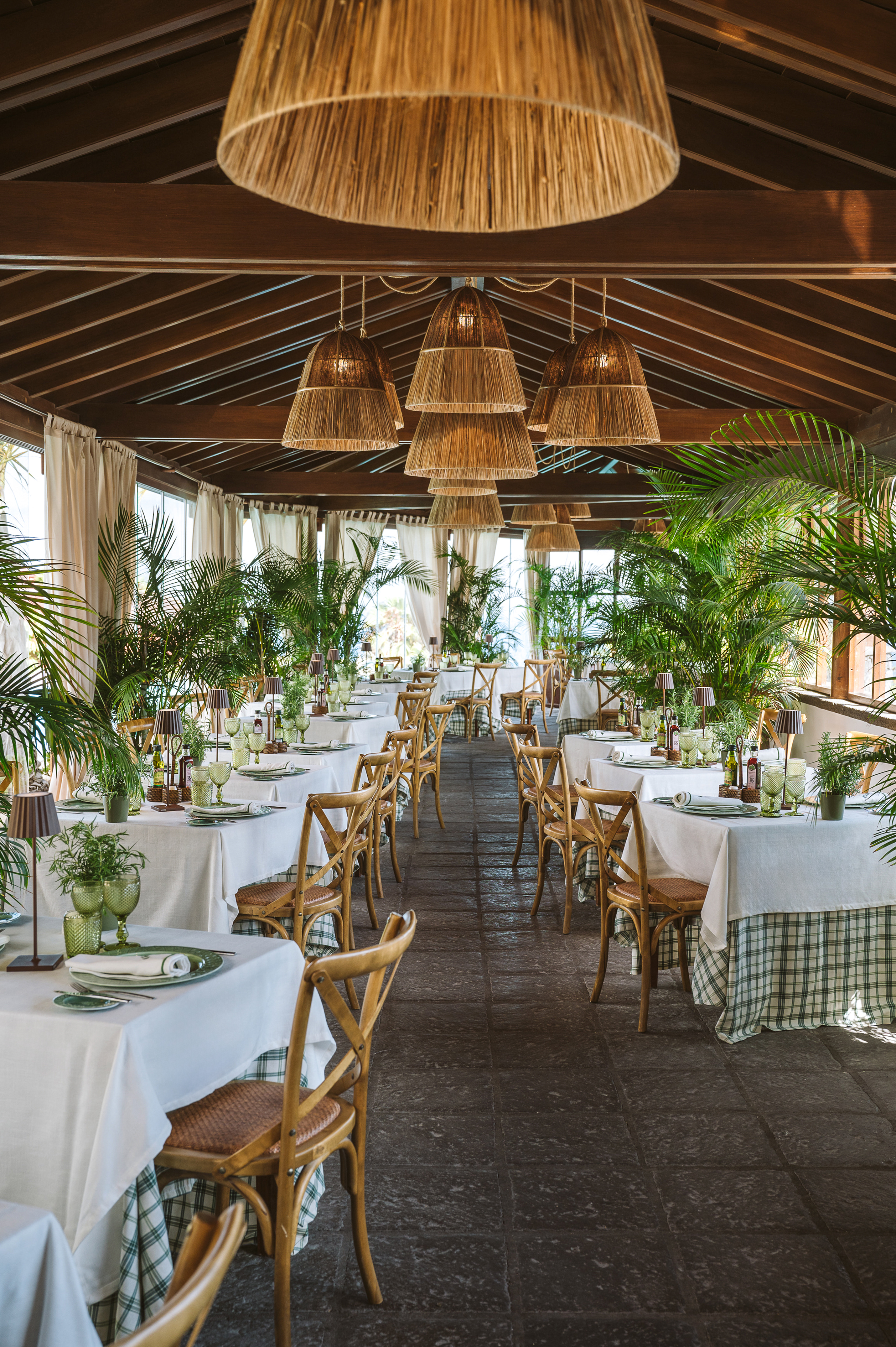 Tropical dining room with palm trees, set tables, wooden chairs, and woven pendant lights.