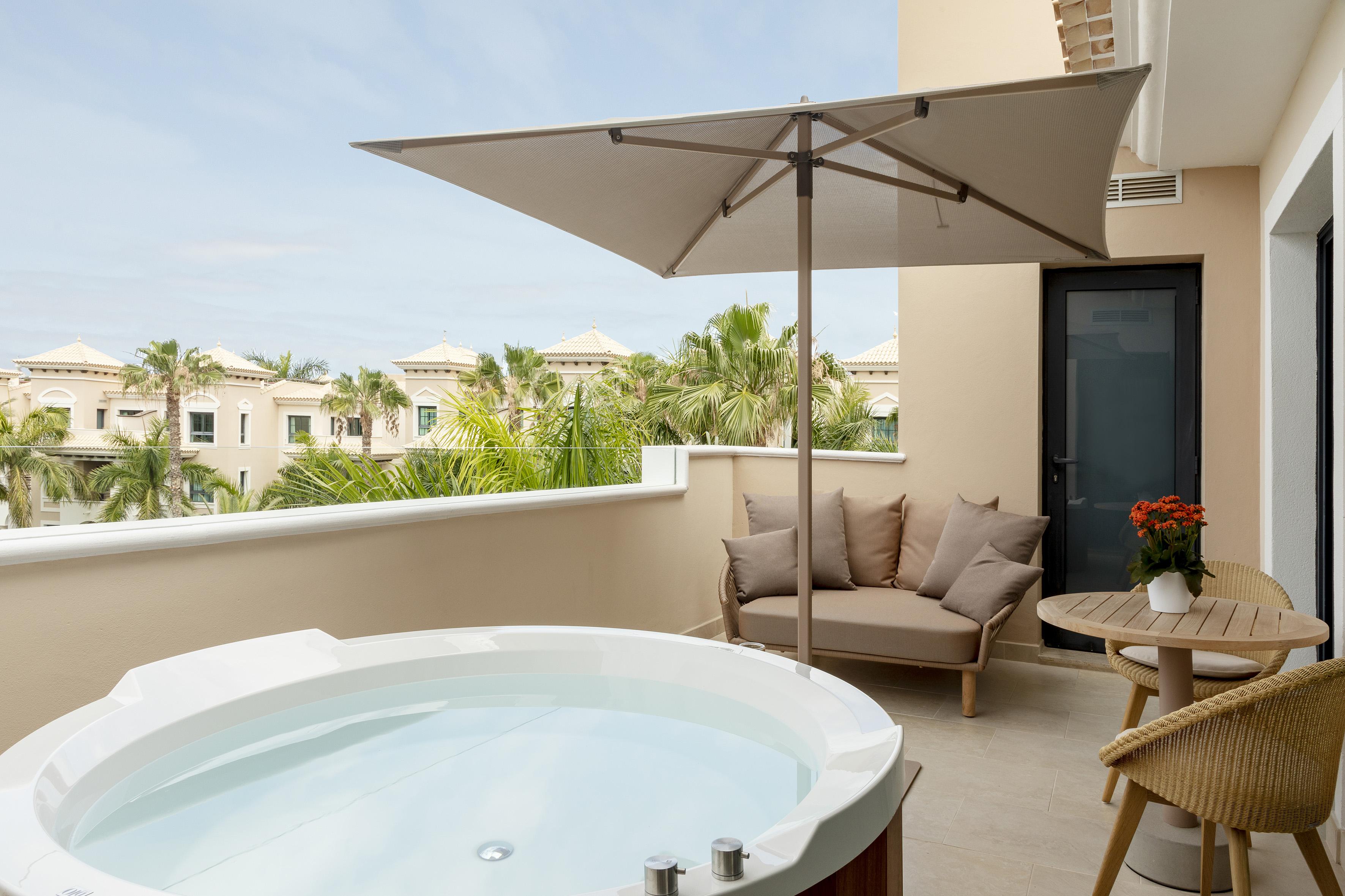 Resort balcony with hot tub, patio furniture, sun umbrella, and palm trees.