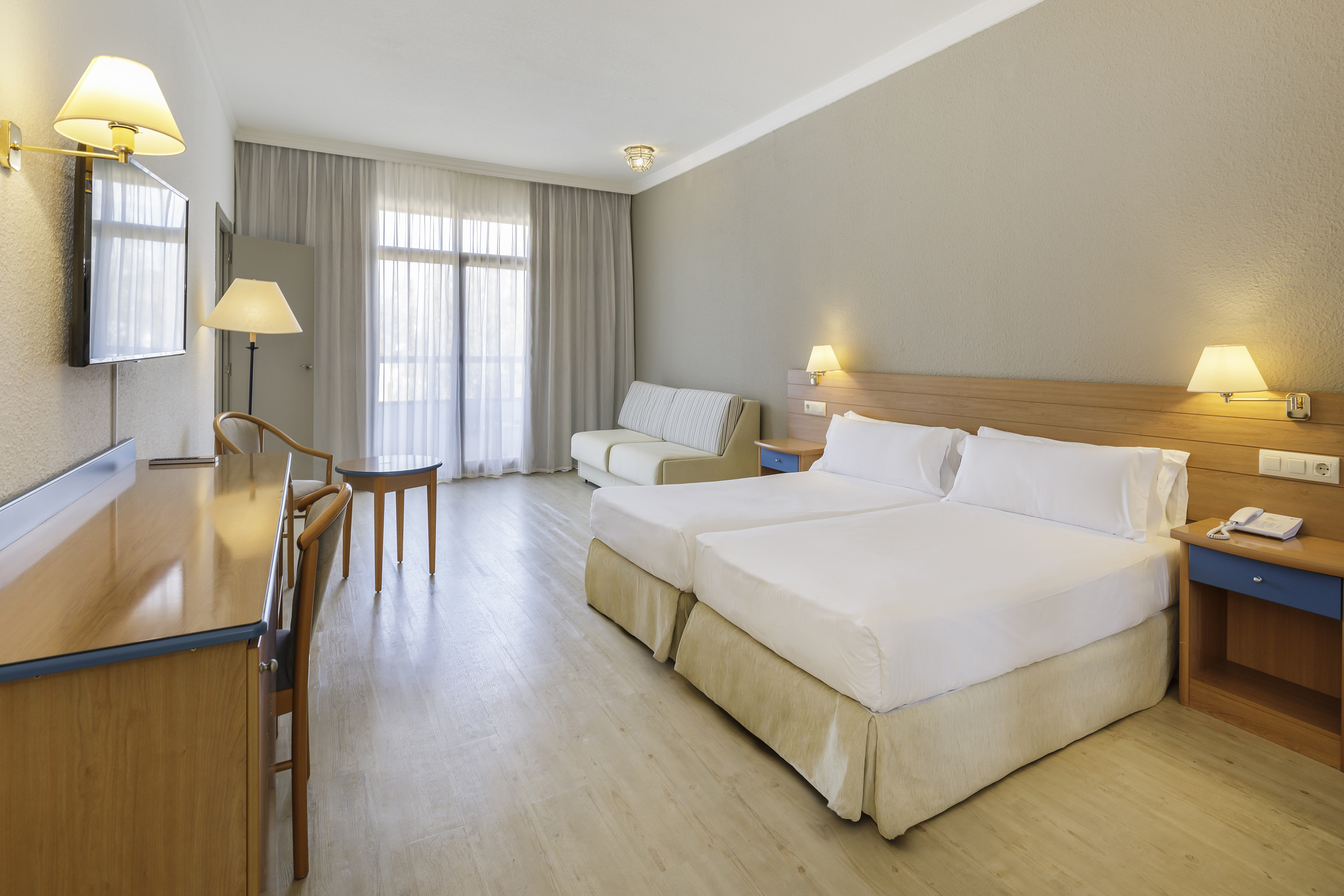 Bright hotel room with two beds, sofa, desk, and large window.