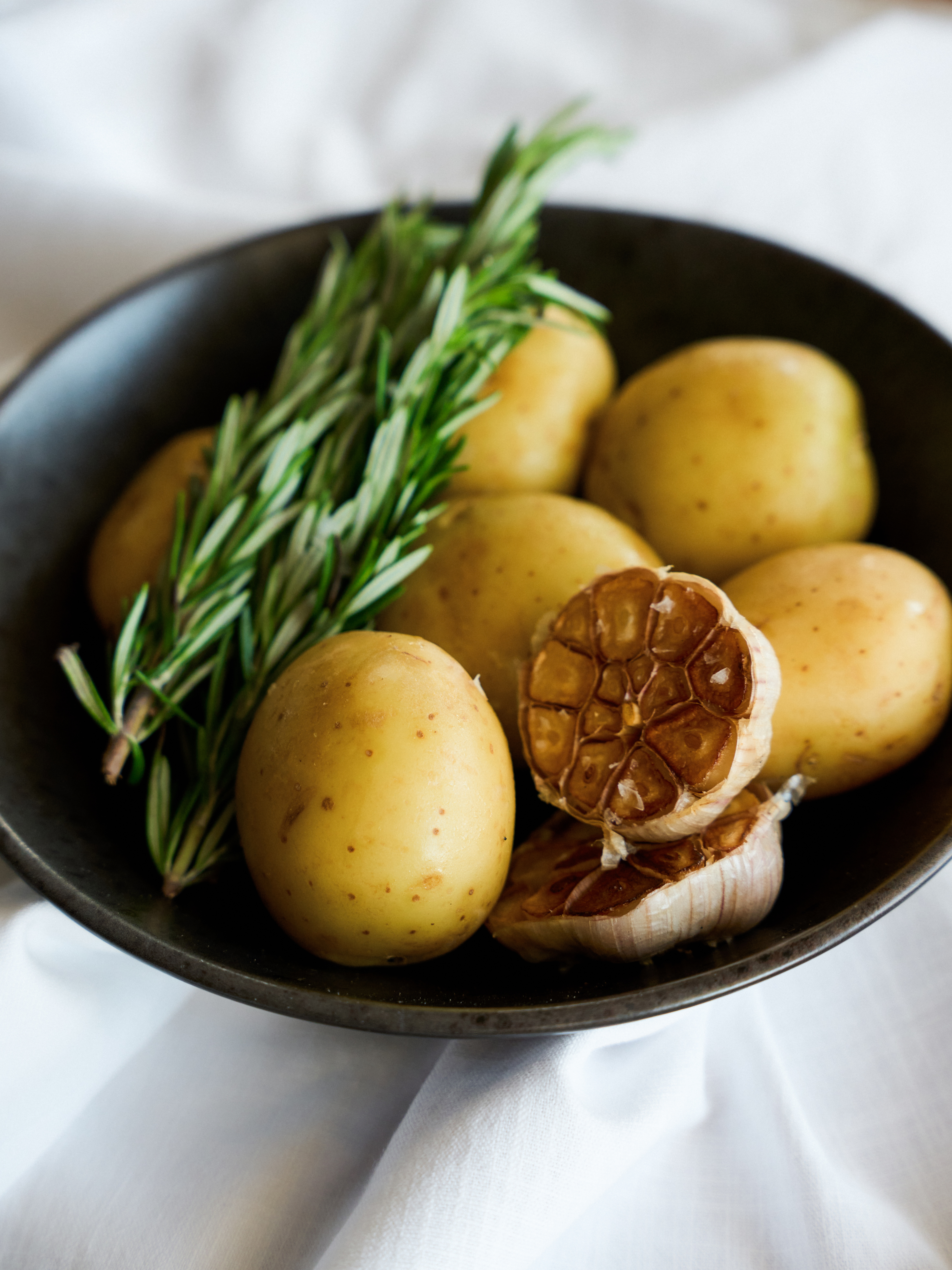 Baby potatoes, fresh rosemary, and roasted garlic in a dark bowl.