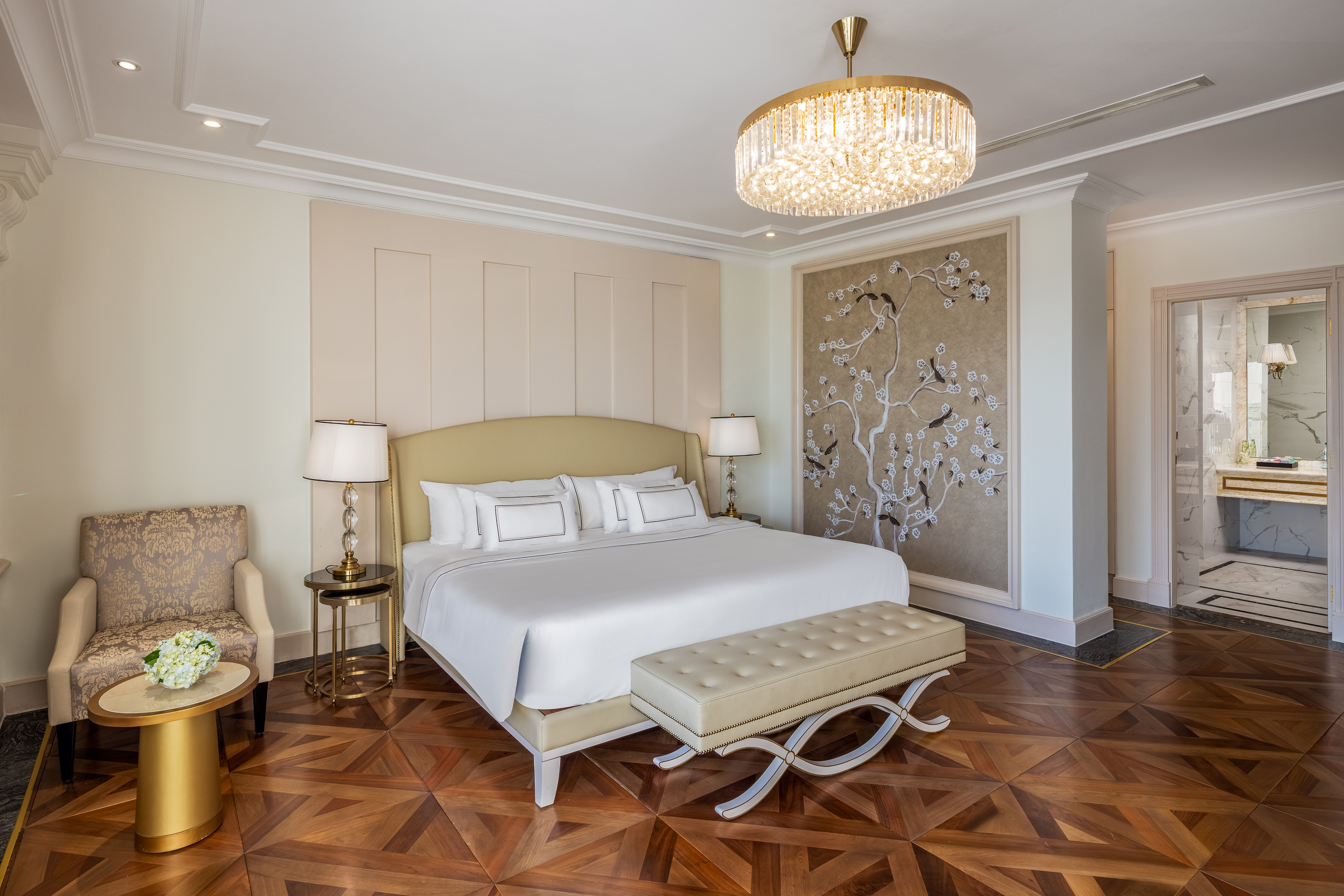 Opulent hotel bedroom: king bed, crystal chandelier, intricate wood floor.