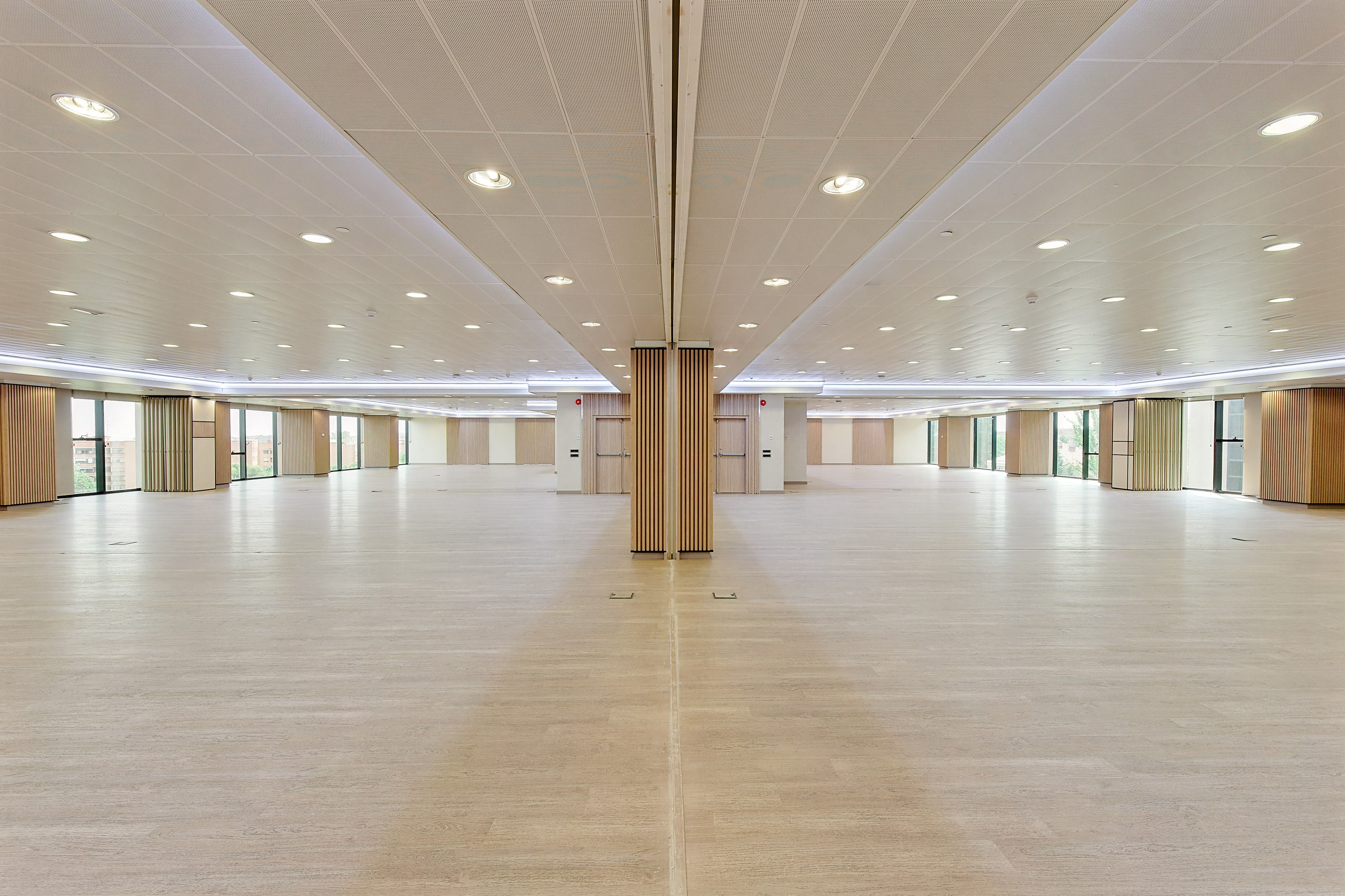 Modern empty event hall with light wood floor, bright ceiling, and windows.