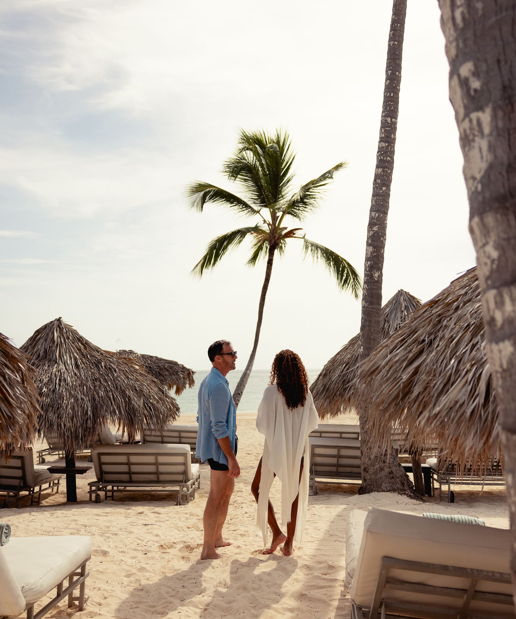 Couple on tropical beach with palm trees, straw umbrellas, and ocean.