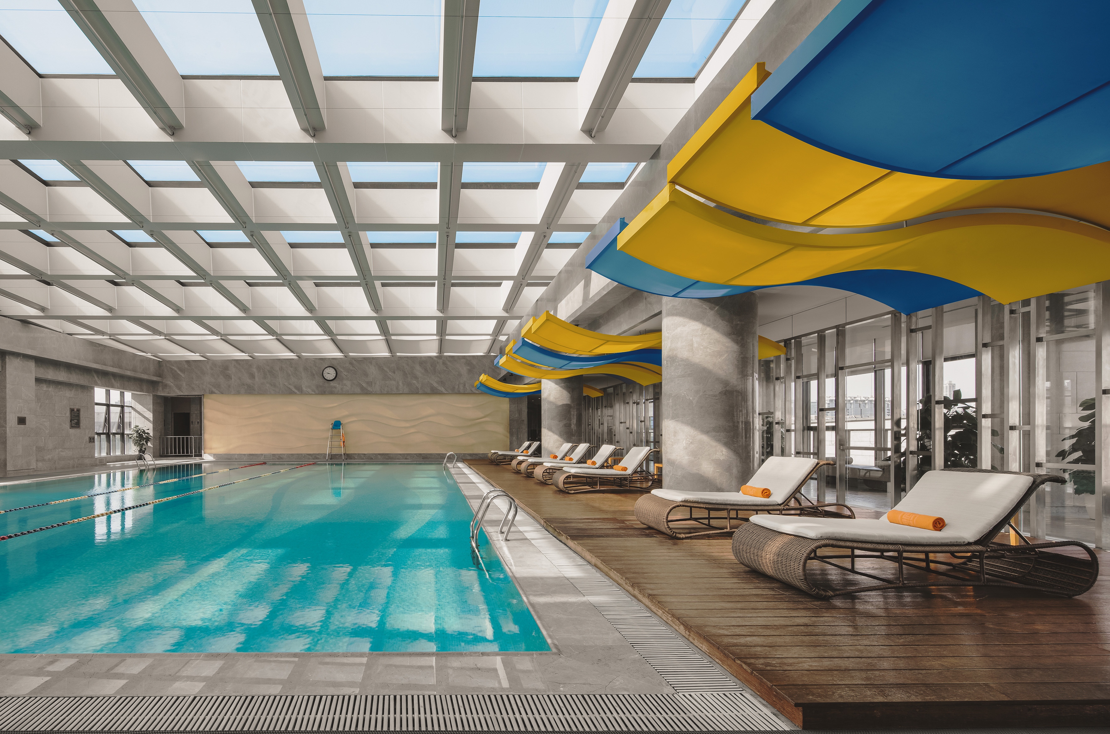 Modern indoor swimming pool with lounge chairs, skylight, and blue/yellow decor.