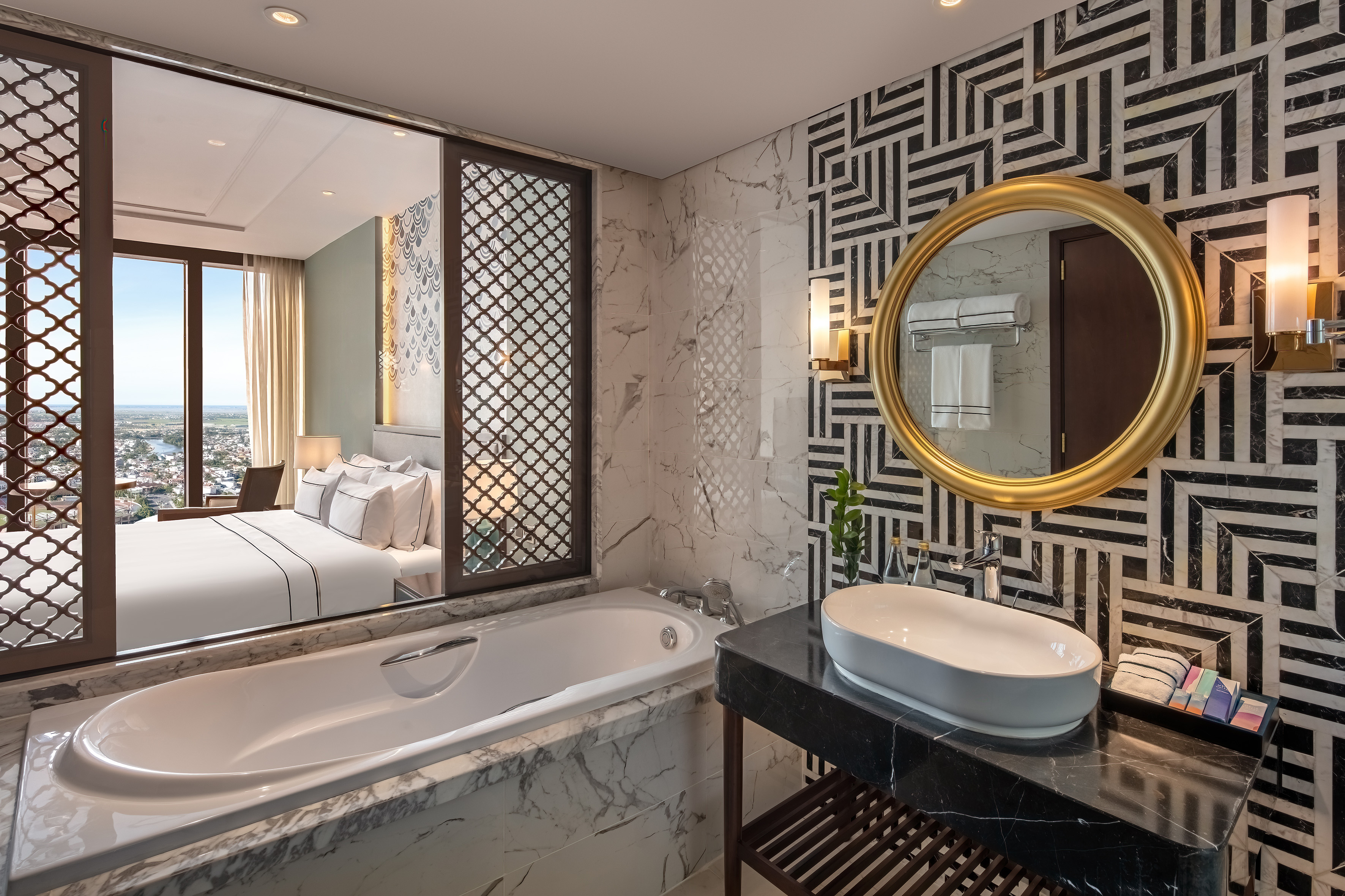 Hotel bathroom: marble bathtub, gold mirror, geometric wall, adjacent bedroom.