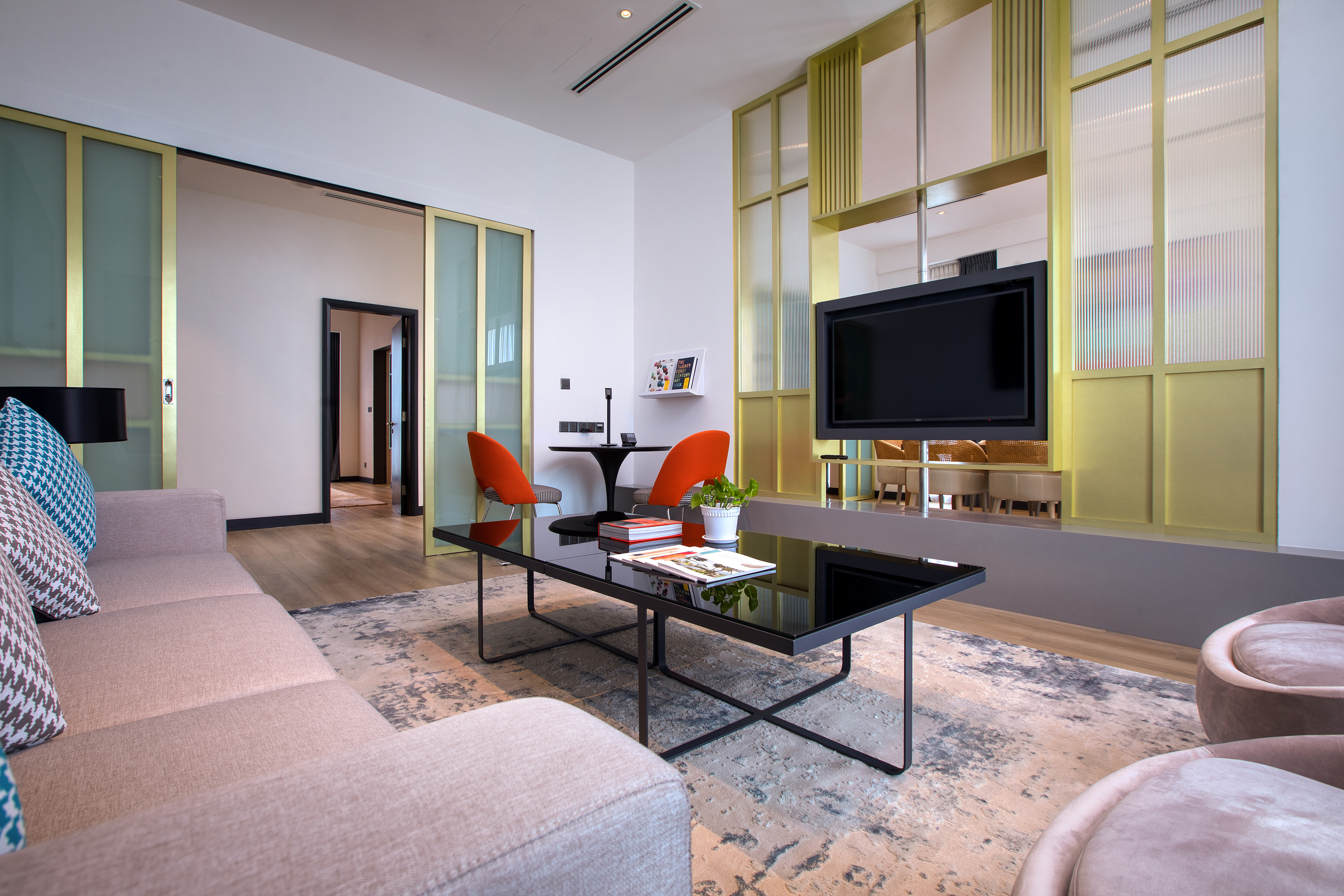 Modern hotel suite with sofa, coffee table, orange chairs, and TV.