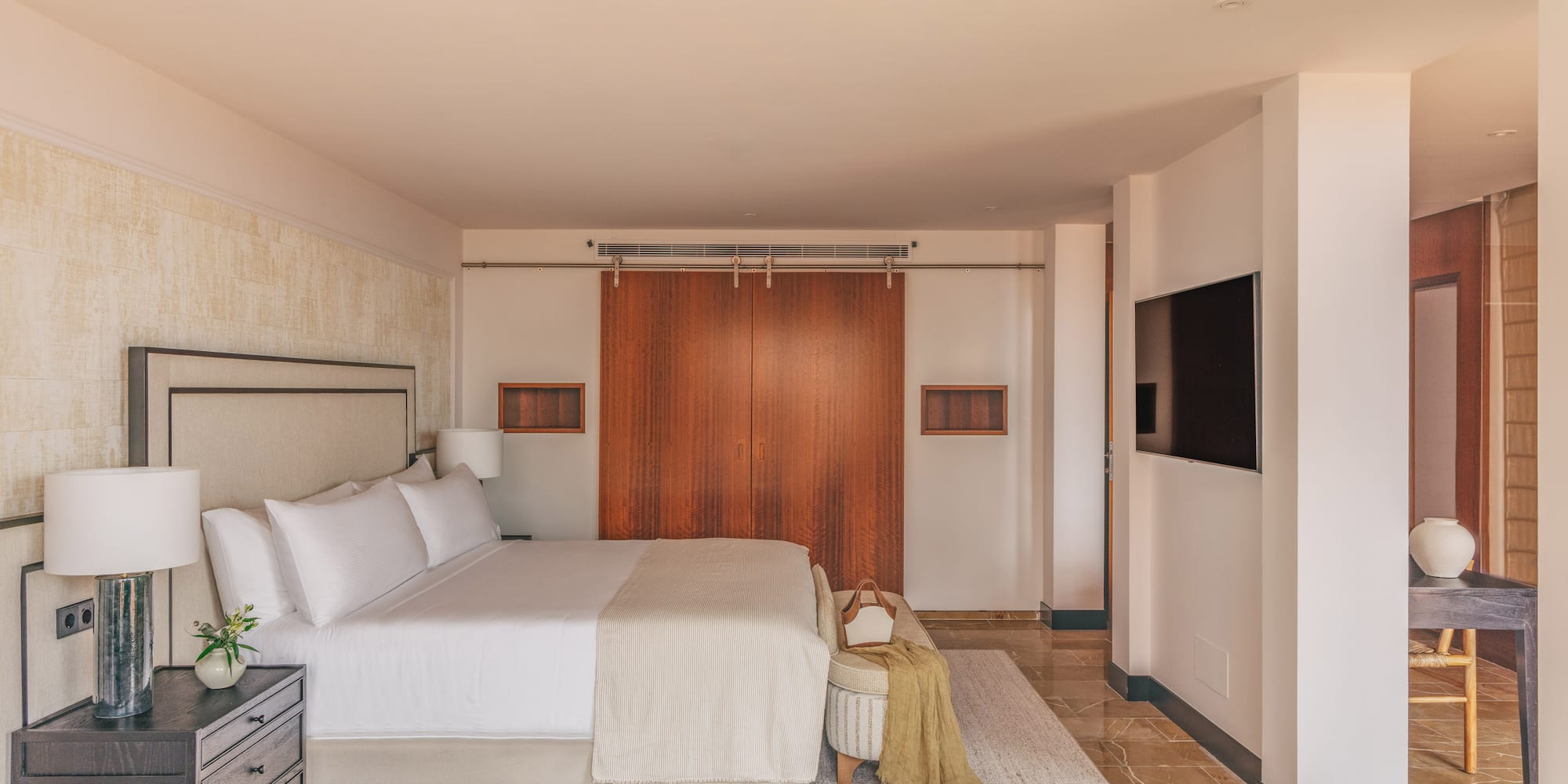 Luxurious modern bedroom with king bed, wooden doors, wall-mounted TV, and marble floor.