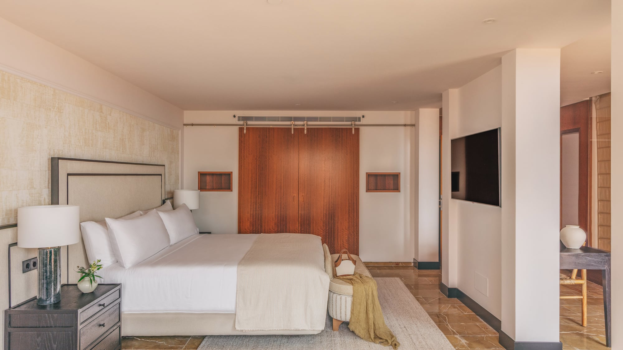 Luxurious modern bedroom with king bed, wooden doors, wall-mounted TV, and marble floor.