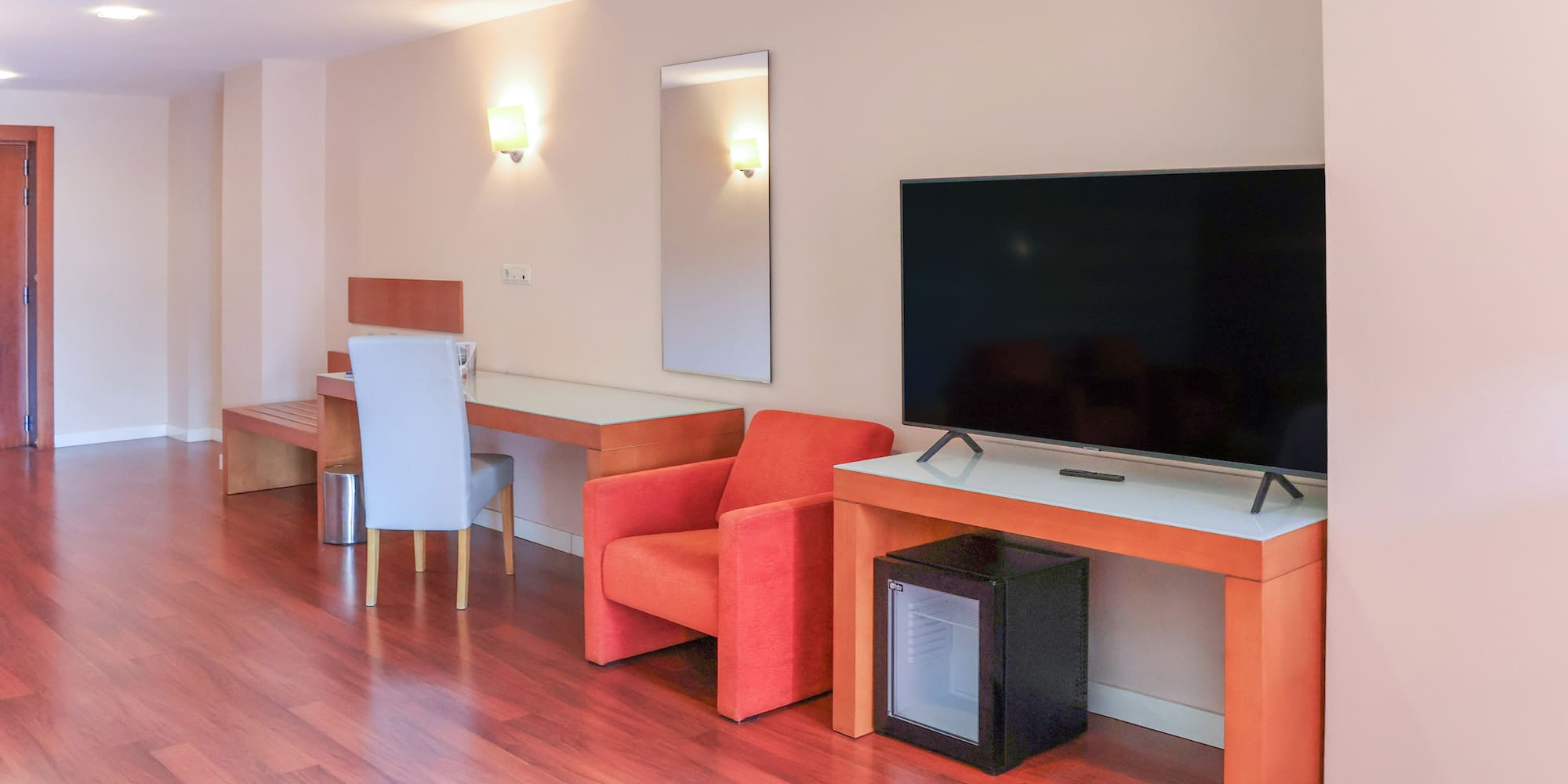 Modern hotel room with TV, desk, orange armchair, mini-fridge, and wood floor.
