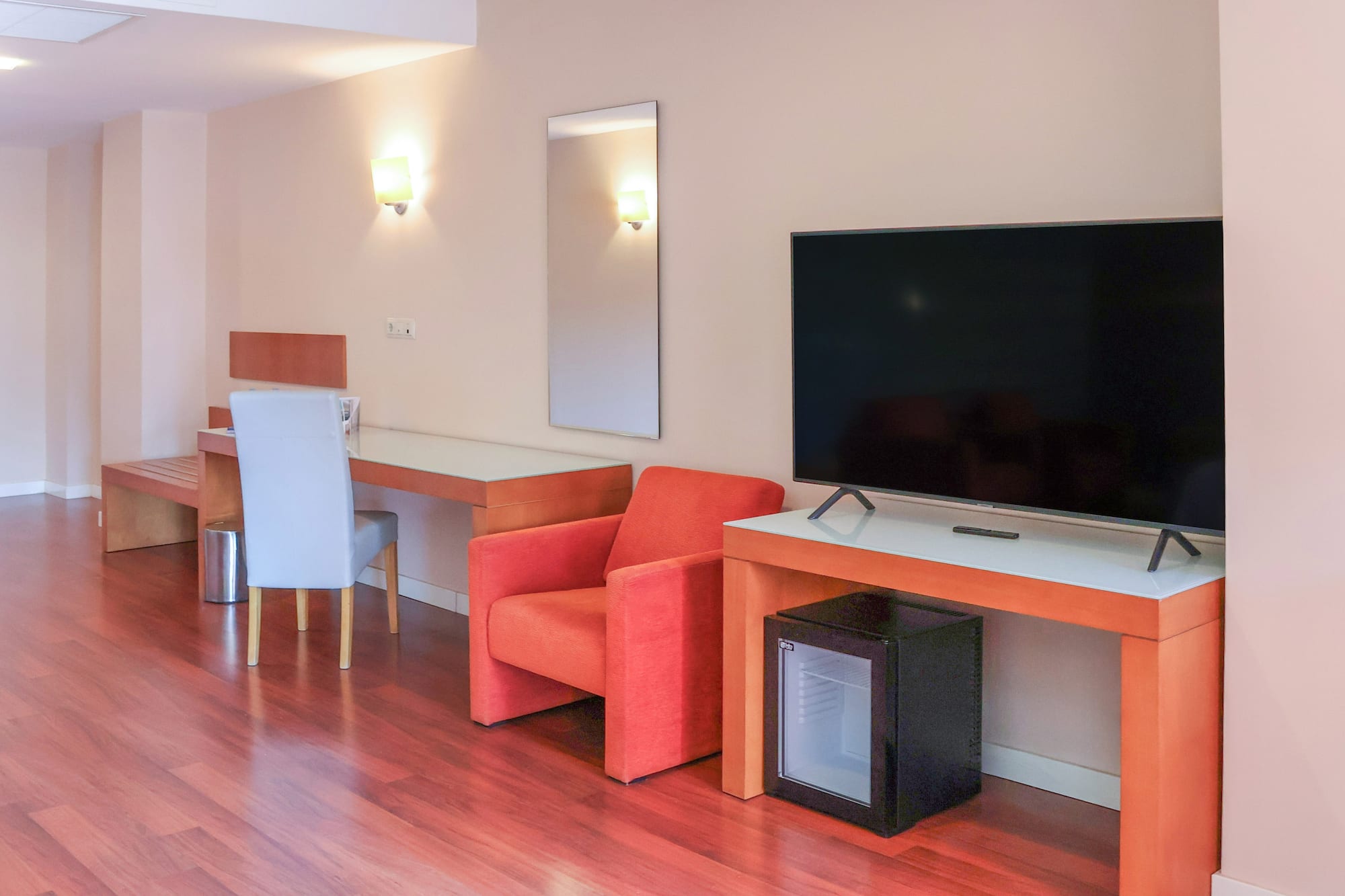 Modern hotel room with TV, desk, orange armchair, mini-fridge, and wood floor.