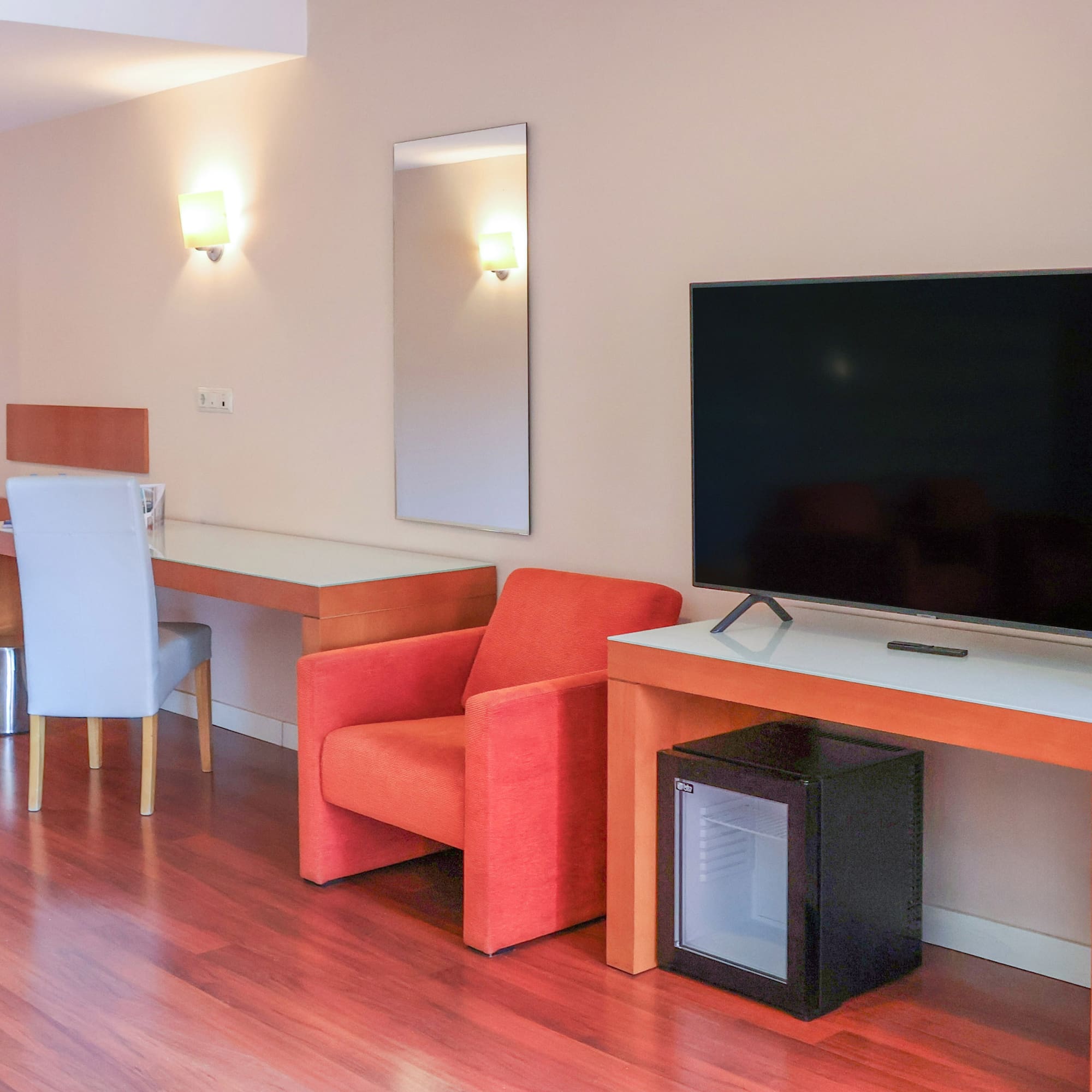 Modern hotel room with TV, desk, orange armchair, mini-fridge, and wood floor.