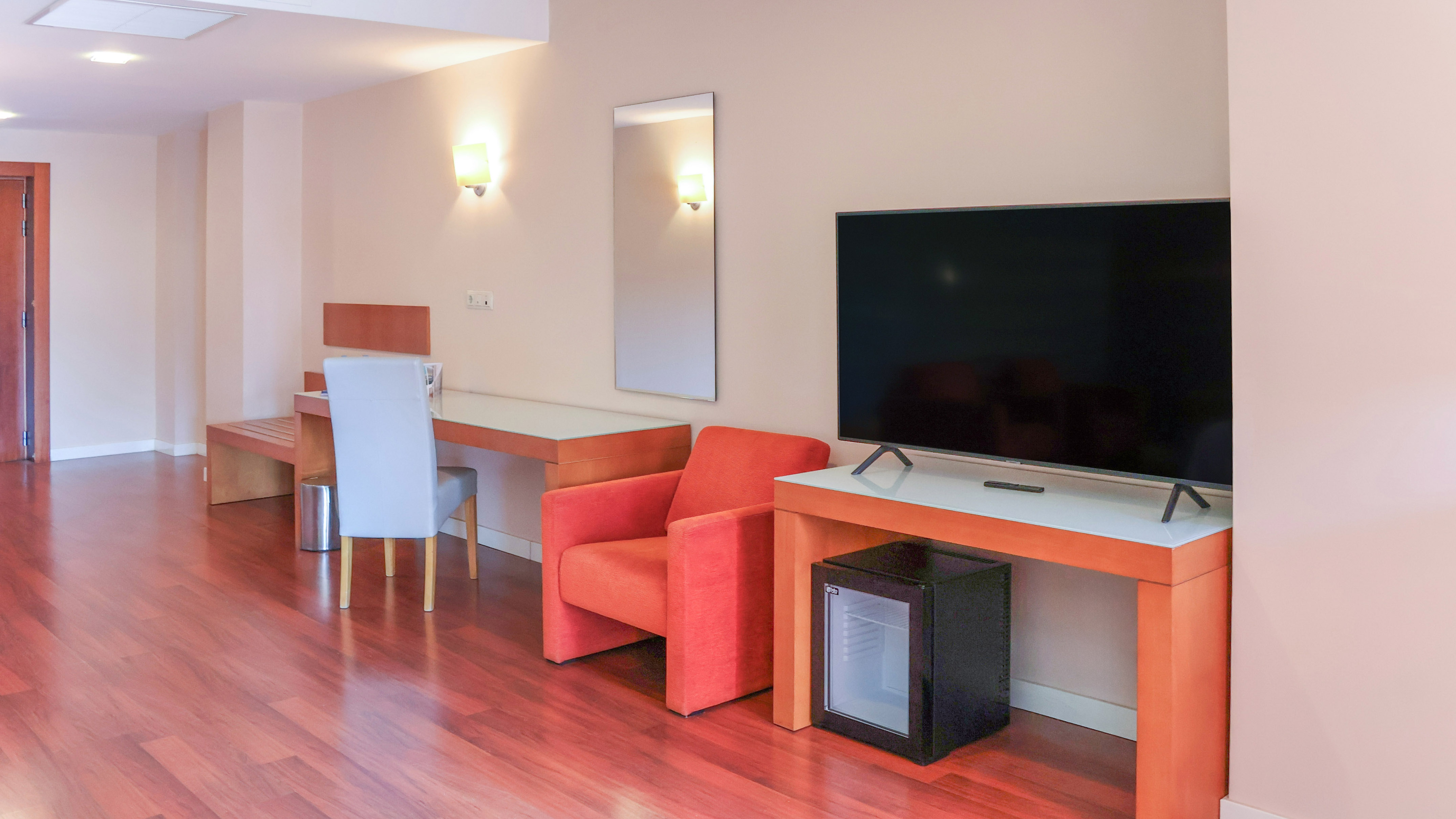 Modern hotel room with TV, desk, orange armchair, mini-fridge, and wood floor.