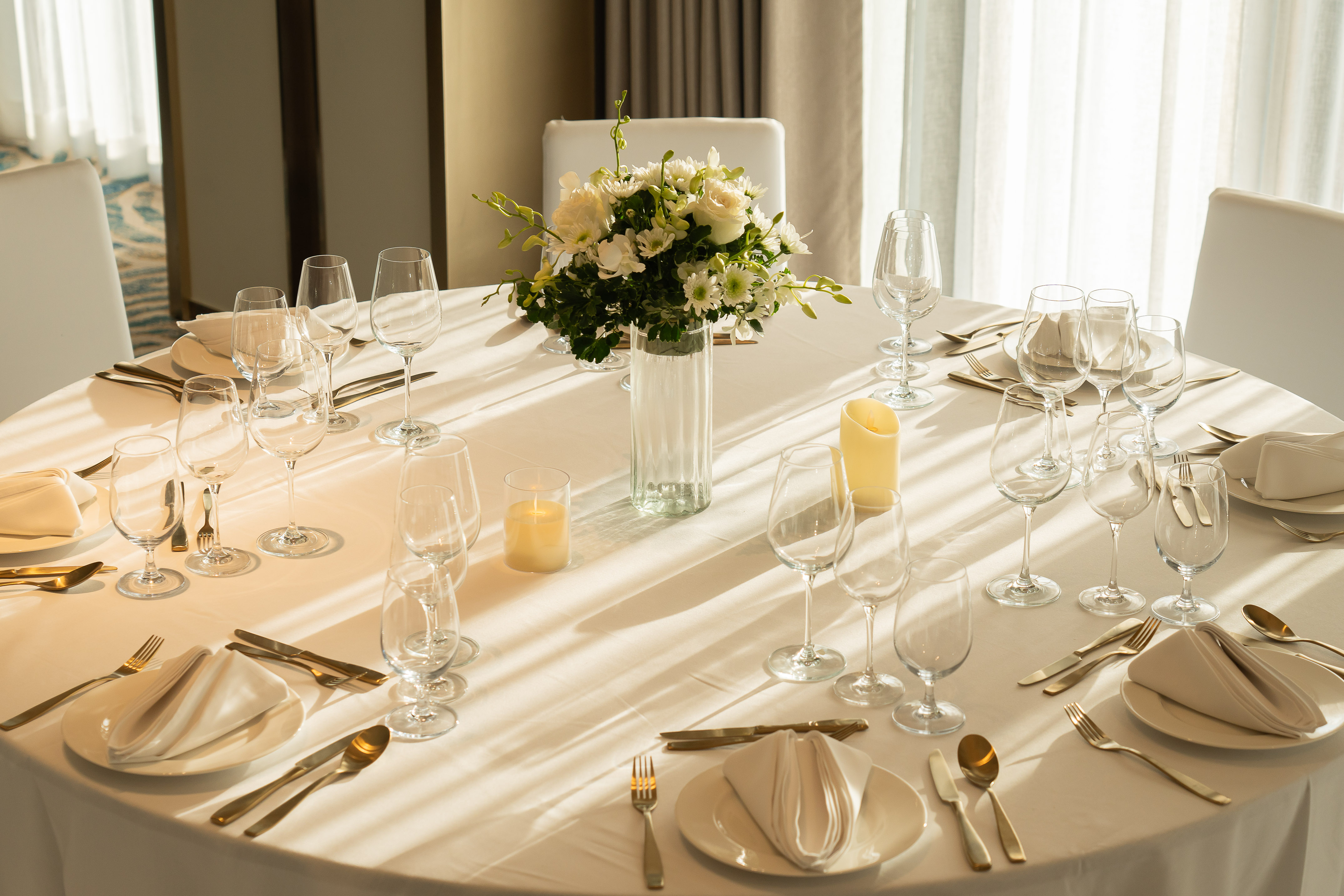 Round event table setting with white flowers, gold cutlery, stemware, and candles. - Nuaan - Meliá Pattaya Hotel, Pattaya