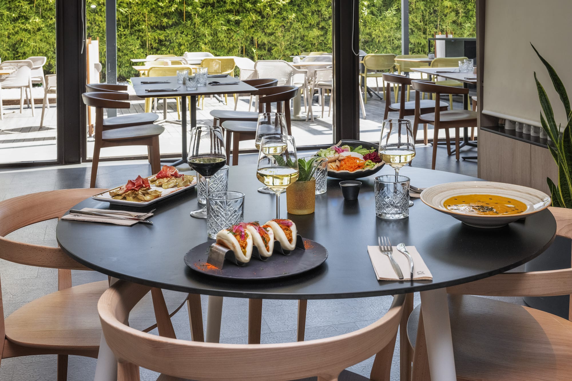 Modern restaurant table with bao buns, soup, wine, and patio view.