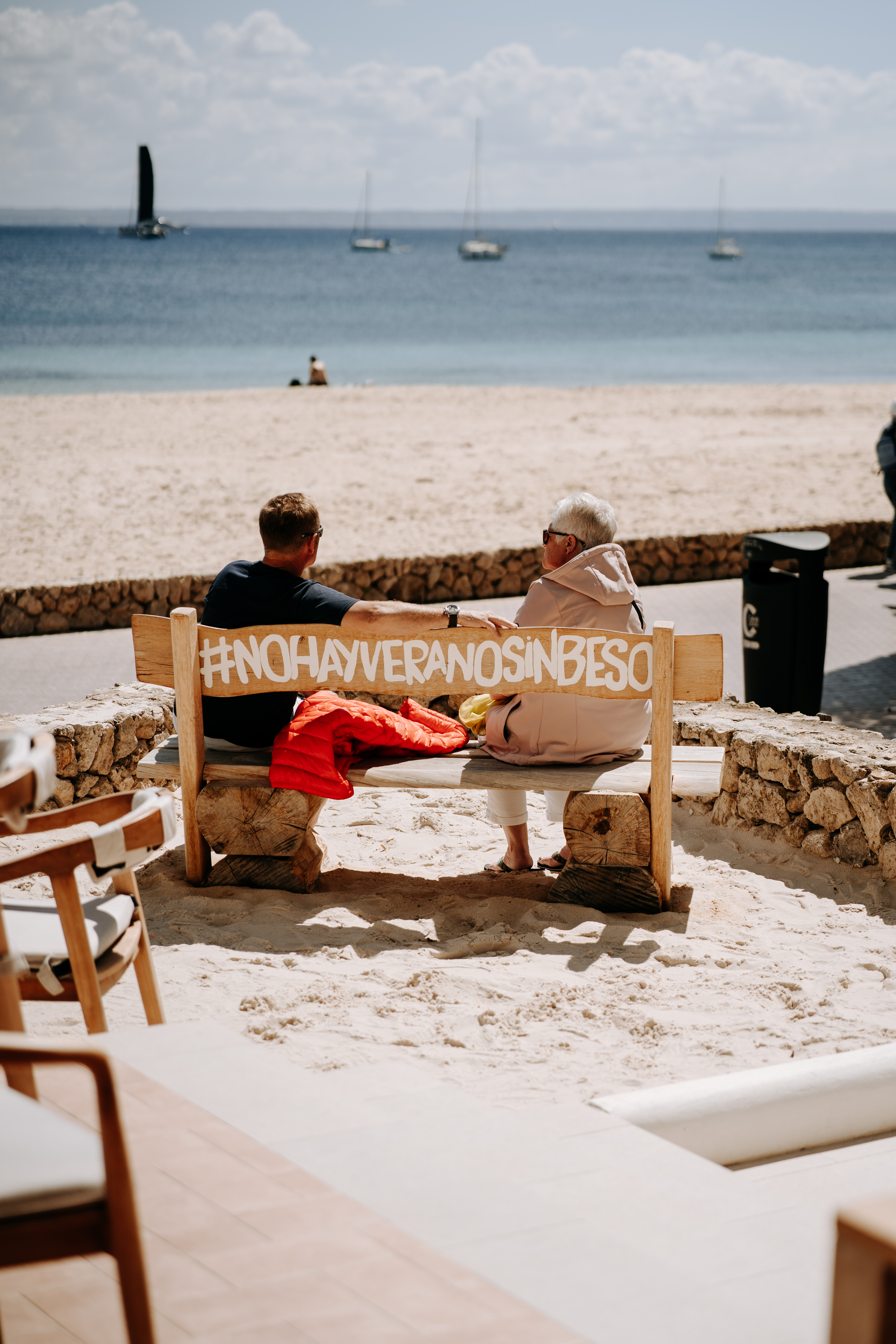 Couple on beach bench, ocean, sailboats; #NOHAYVERANOSINBESO text.
