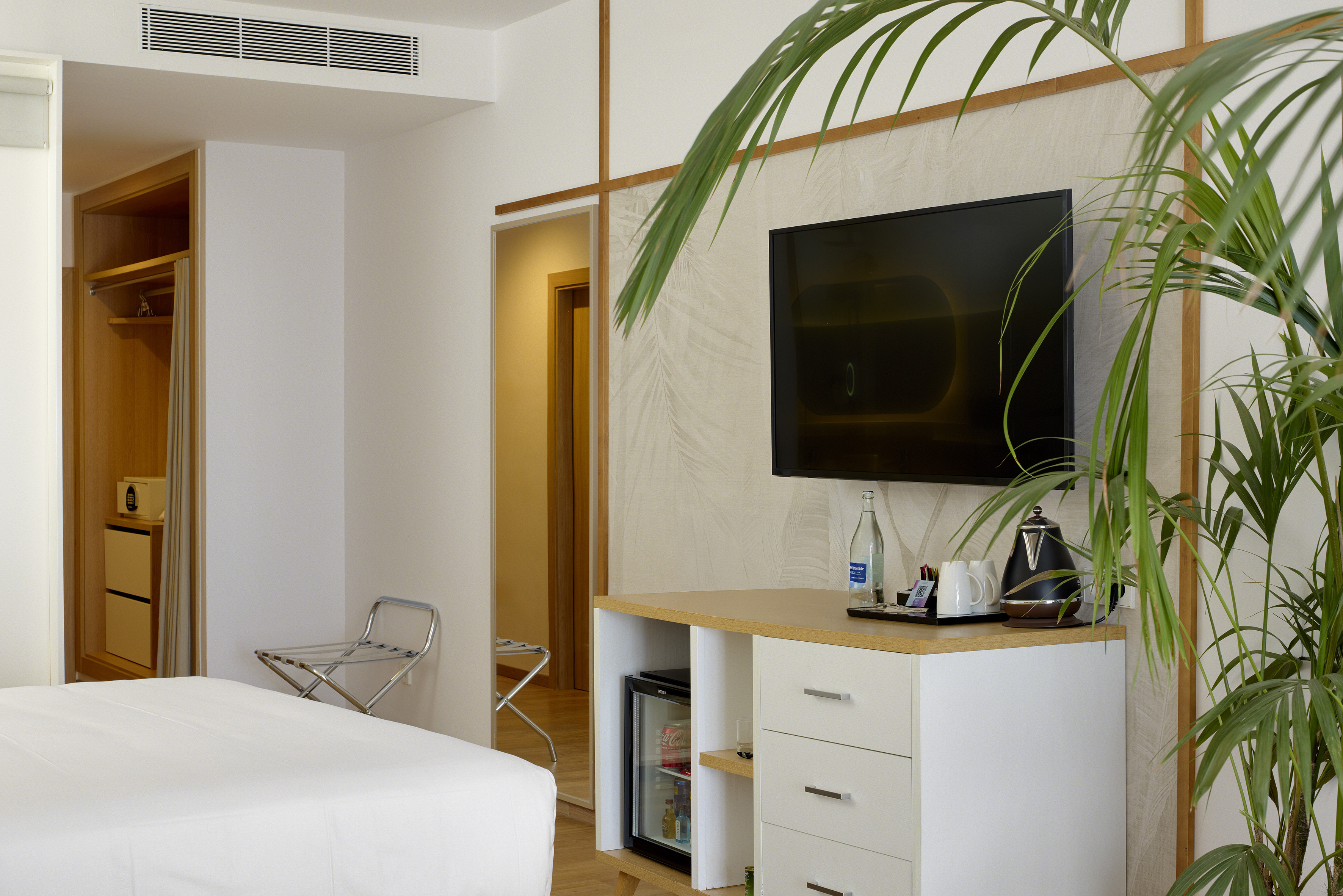 Modern hotel room with TV, mini-fridge, bed, wardrobe, and green plant.