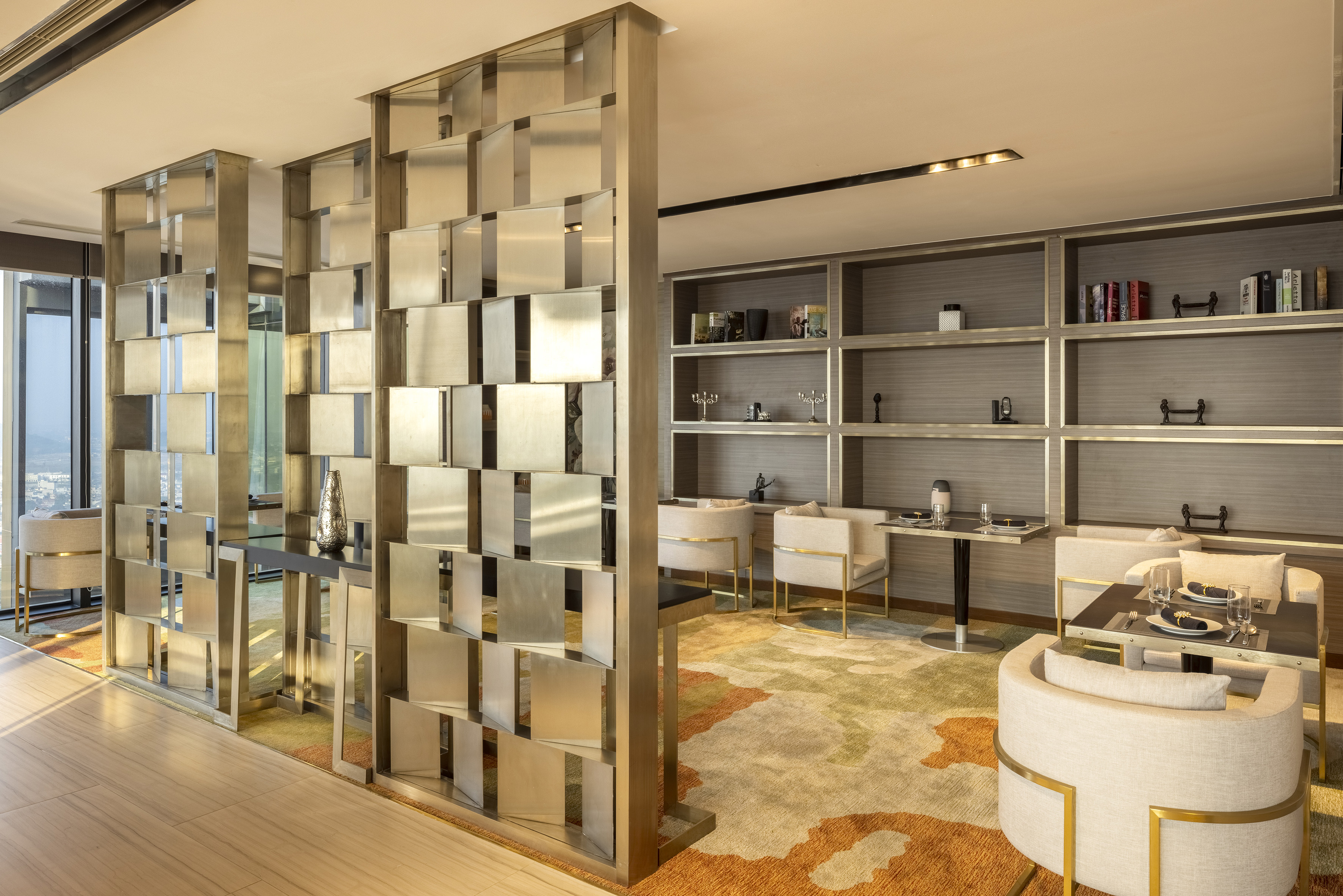 Modern luxury lounge with metallic room divider, cozy seating, patterned rug, and city view.
