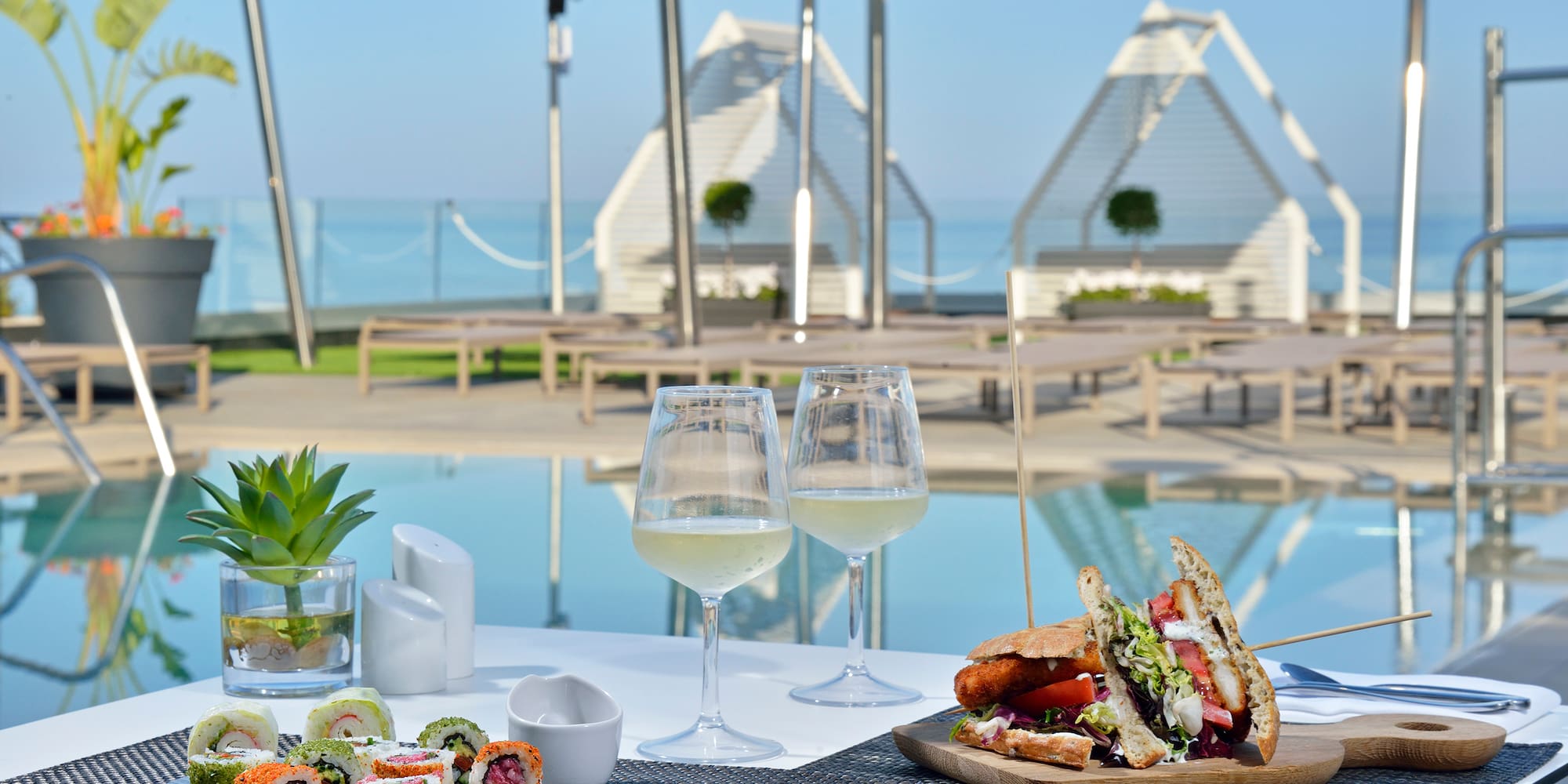 Sushi and sandwich with white wine by a resort infinity pool, ocean view.
