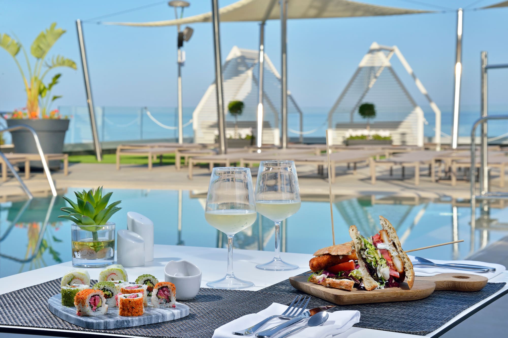 Sushi and sandwich with white wine by a resort infinity pool, ocean view.