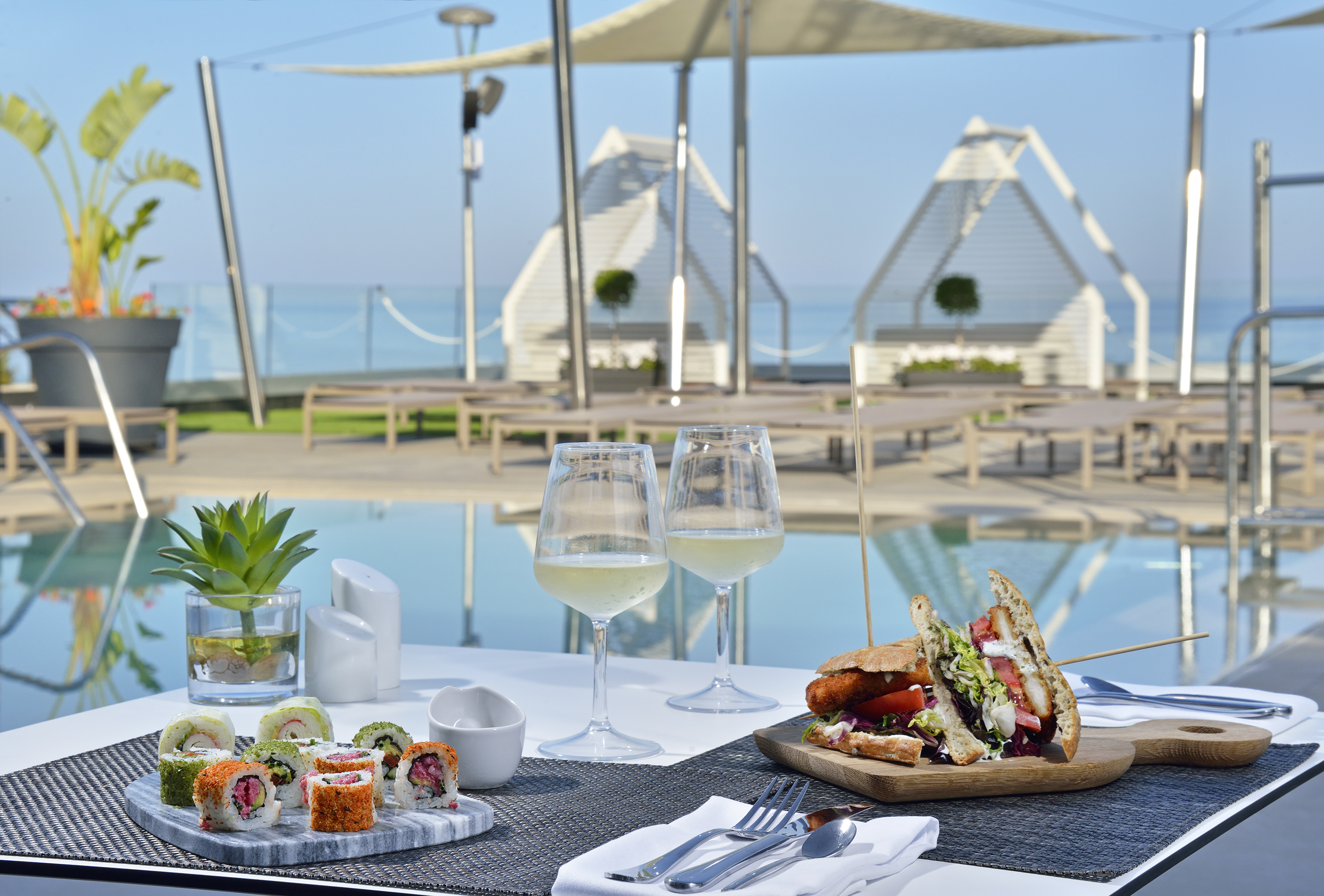 Sushi and sandwich with white wine by a resort infinity pool, ocean view.