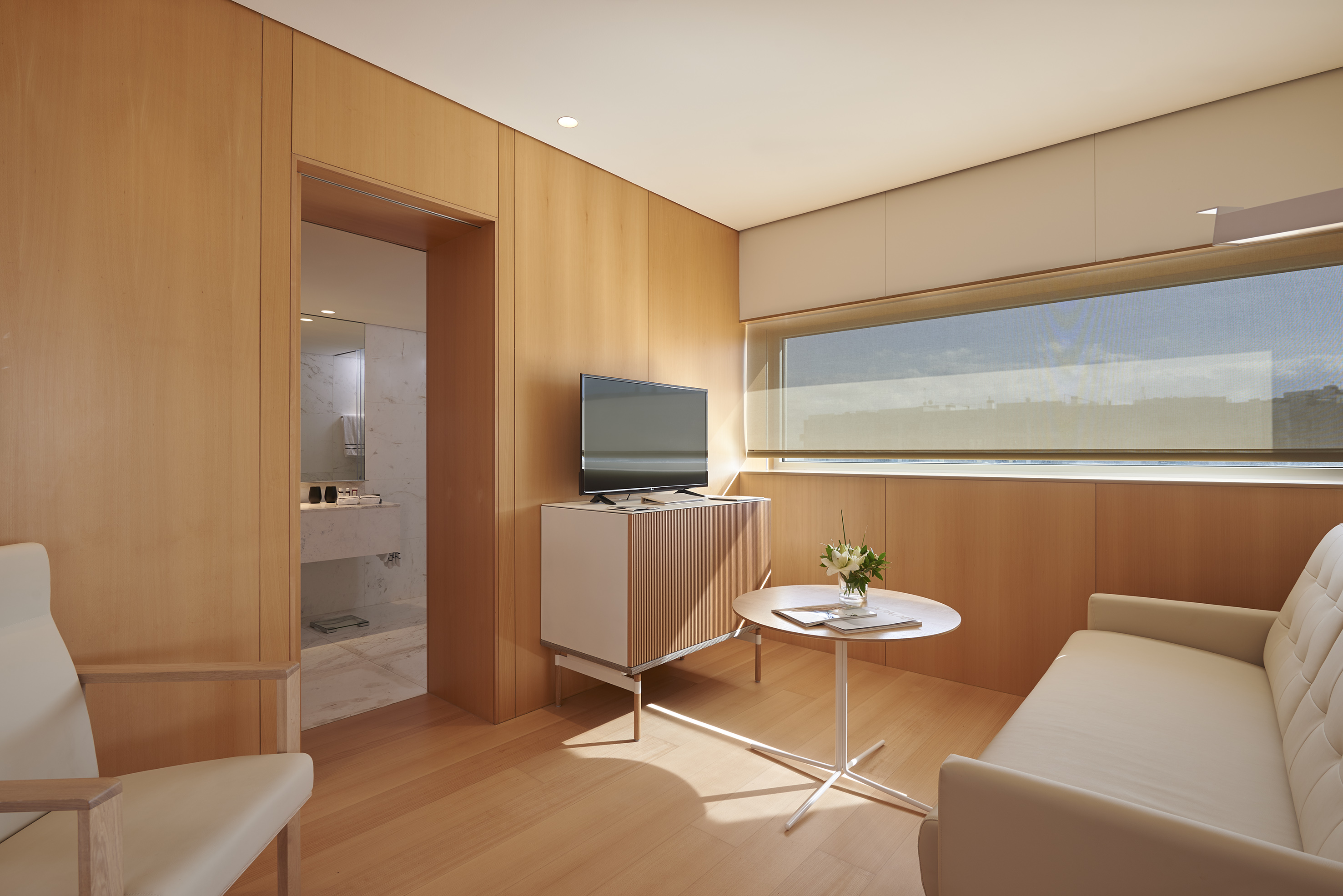 Modern hotel suite with wood paneling, beige seating, TV, and bathroom access.
