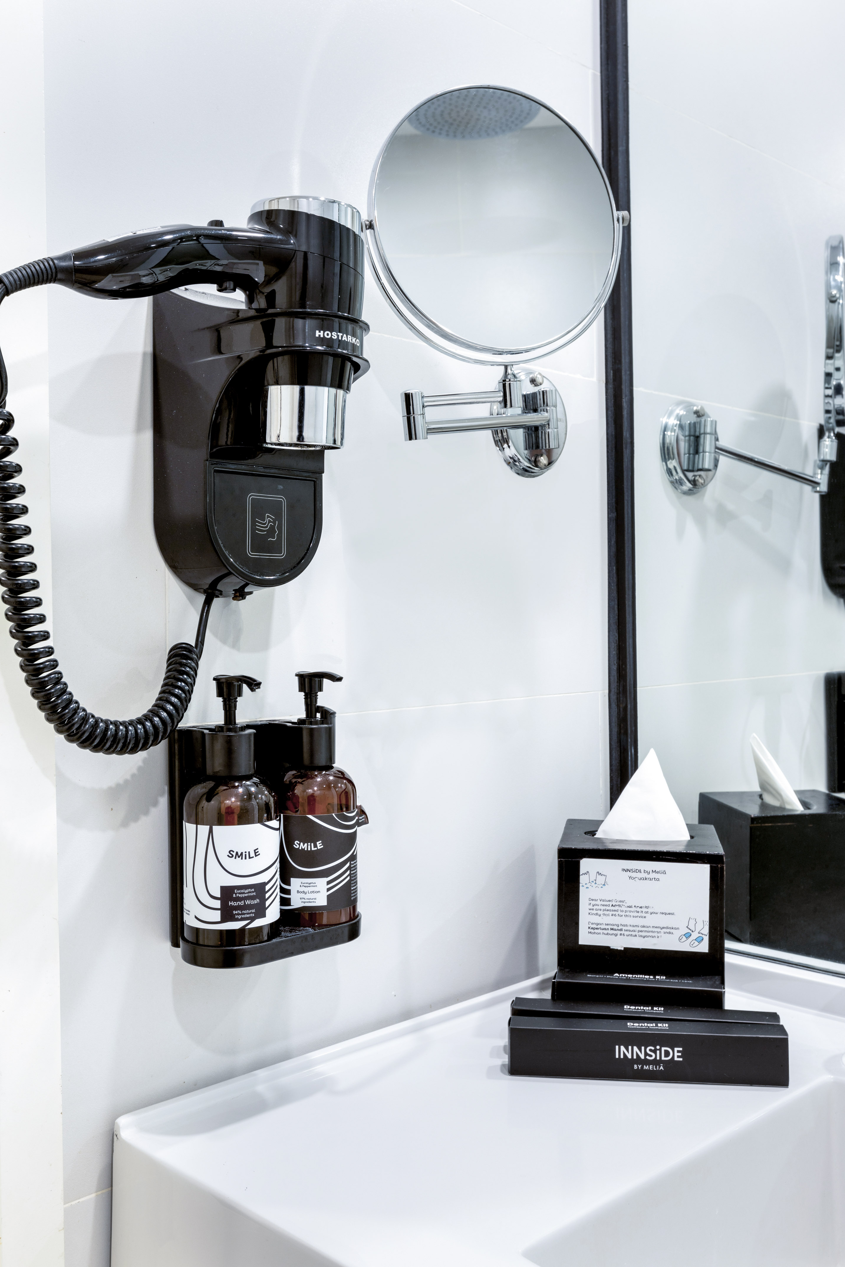 Bathroom vanity with wall-mounted hairdryer, magnifying mirror, and hotel toiletries.