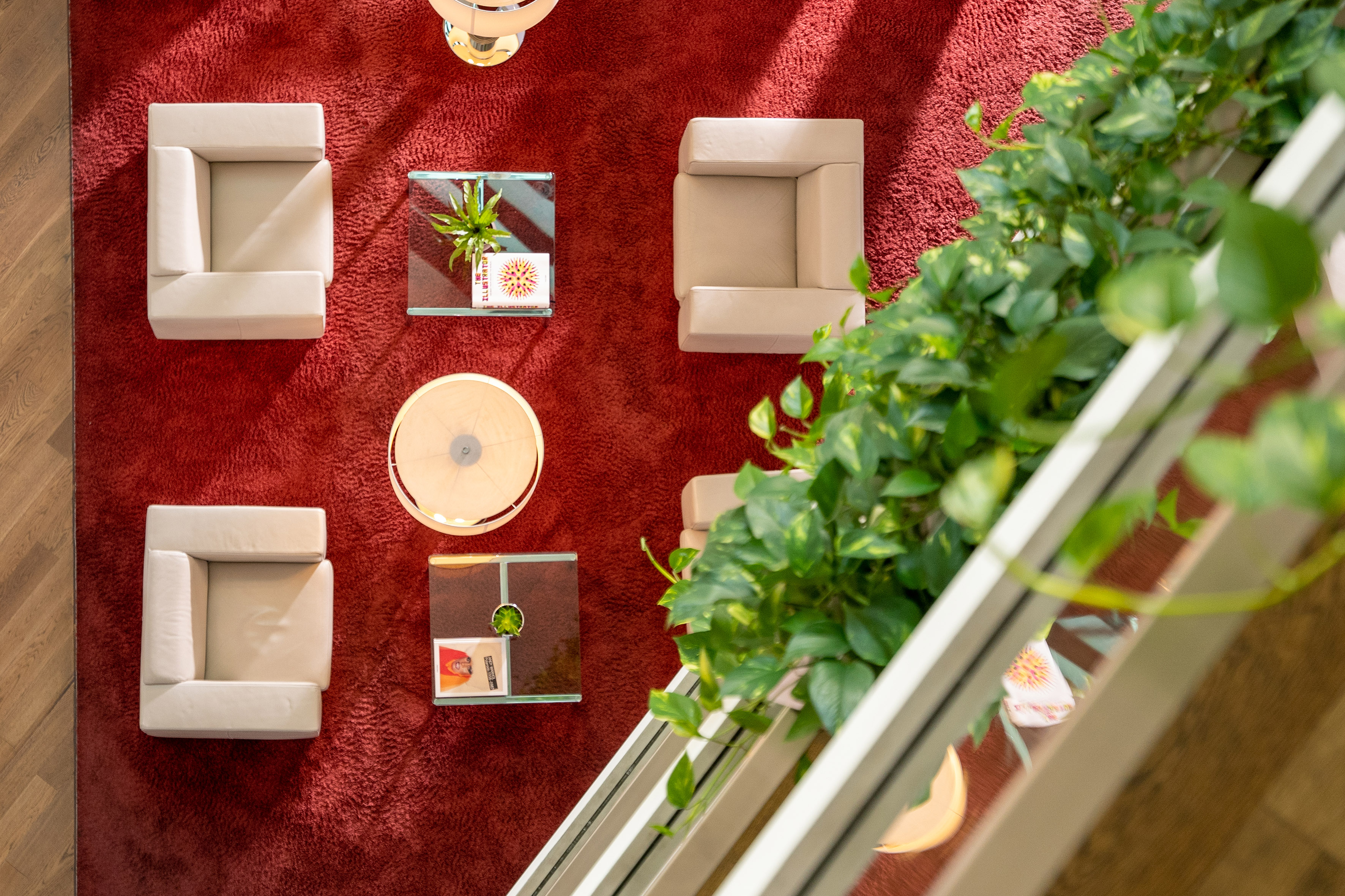 Modern waiting area with beige chairs, glass tables, green plants, red carpet.