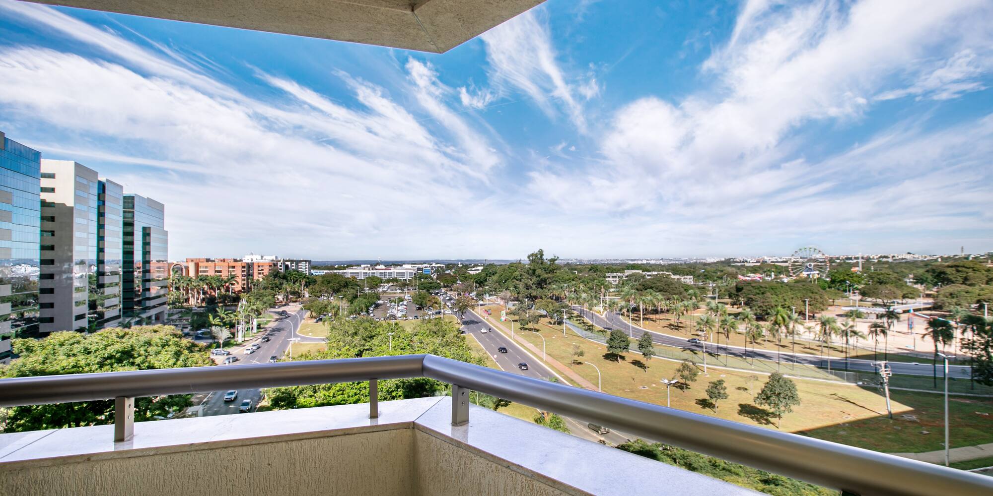 Balcony view: modern buildings, urban streets, green trees, and bright blue sky.