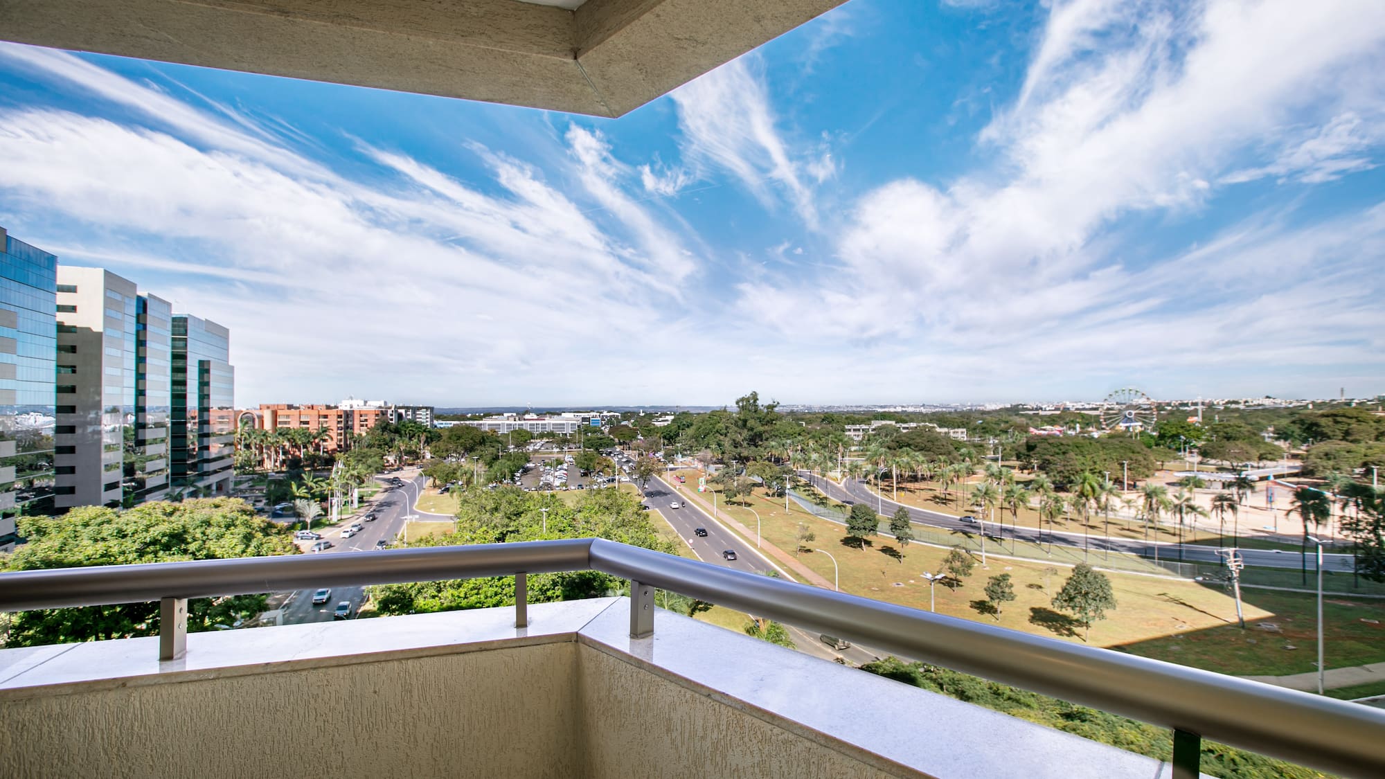 Balcony view: modern buildings, urban streets, green trees, and bright blue sky.