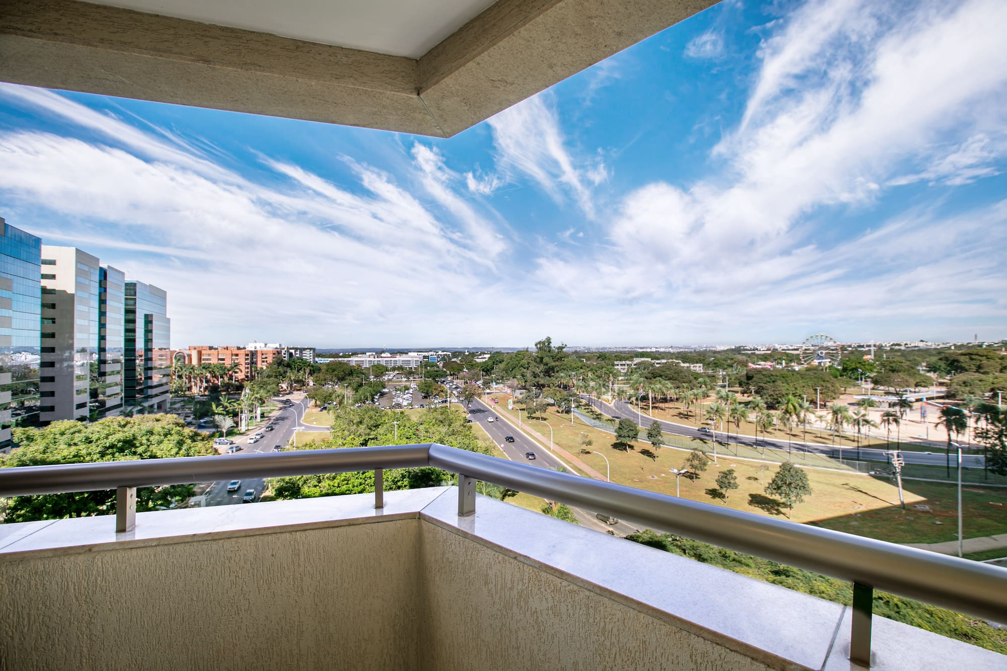 Balcony view: modern buildings, urban streets, green trees, and bright blue sky.